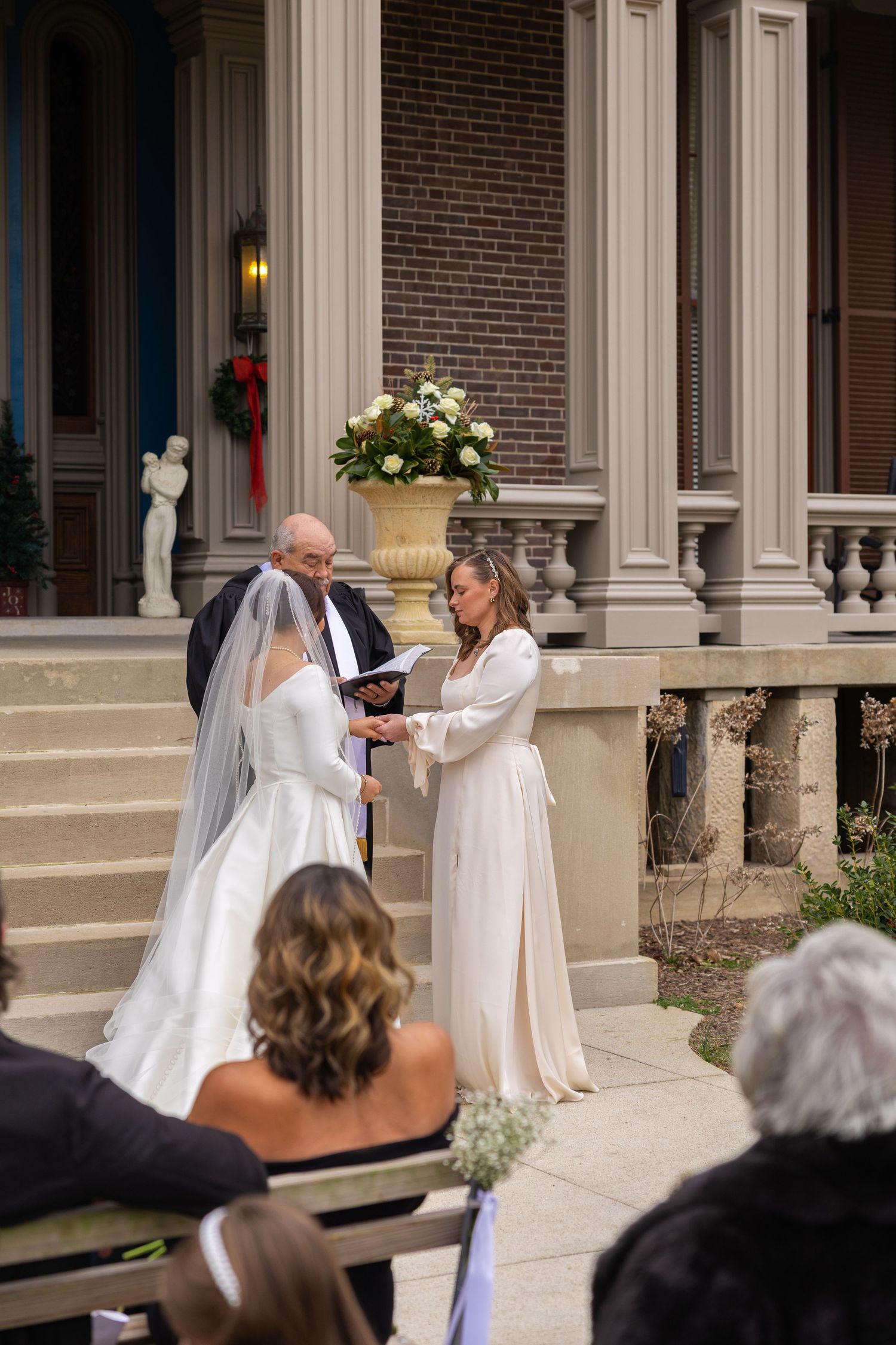Wedding ceremony taking place on the steps of a historic brick building with white columns and guests watching.