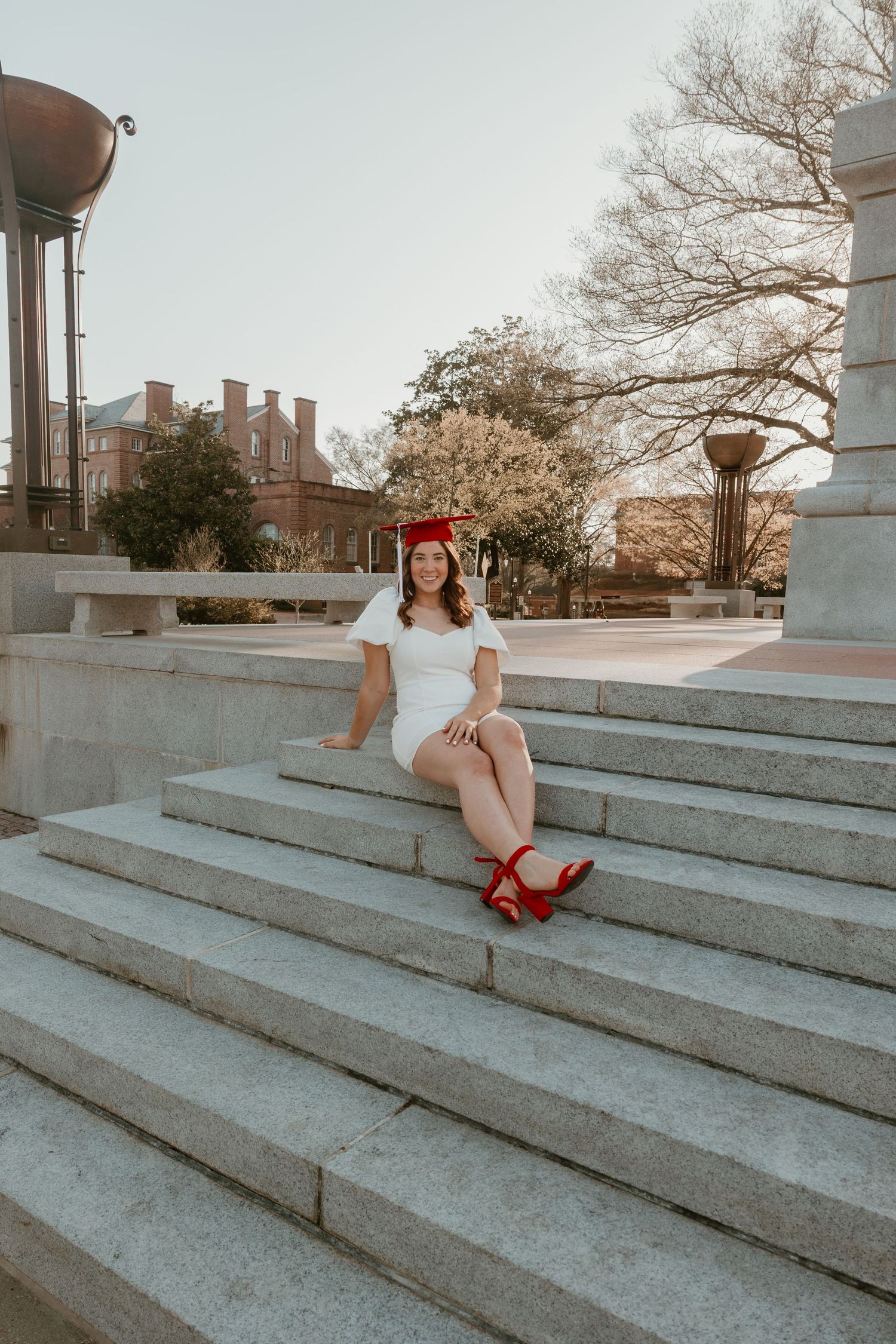 Young woman in white dress and red graduation cap sitting on stone steps, celebrating her graduation outdoors.