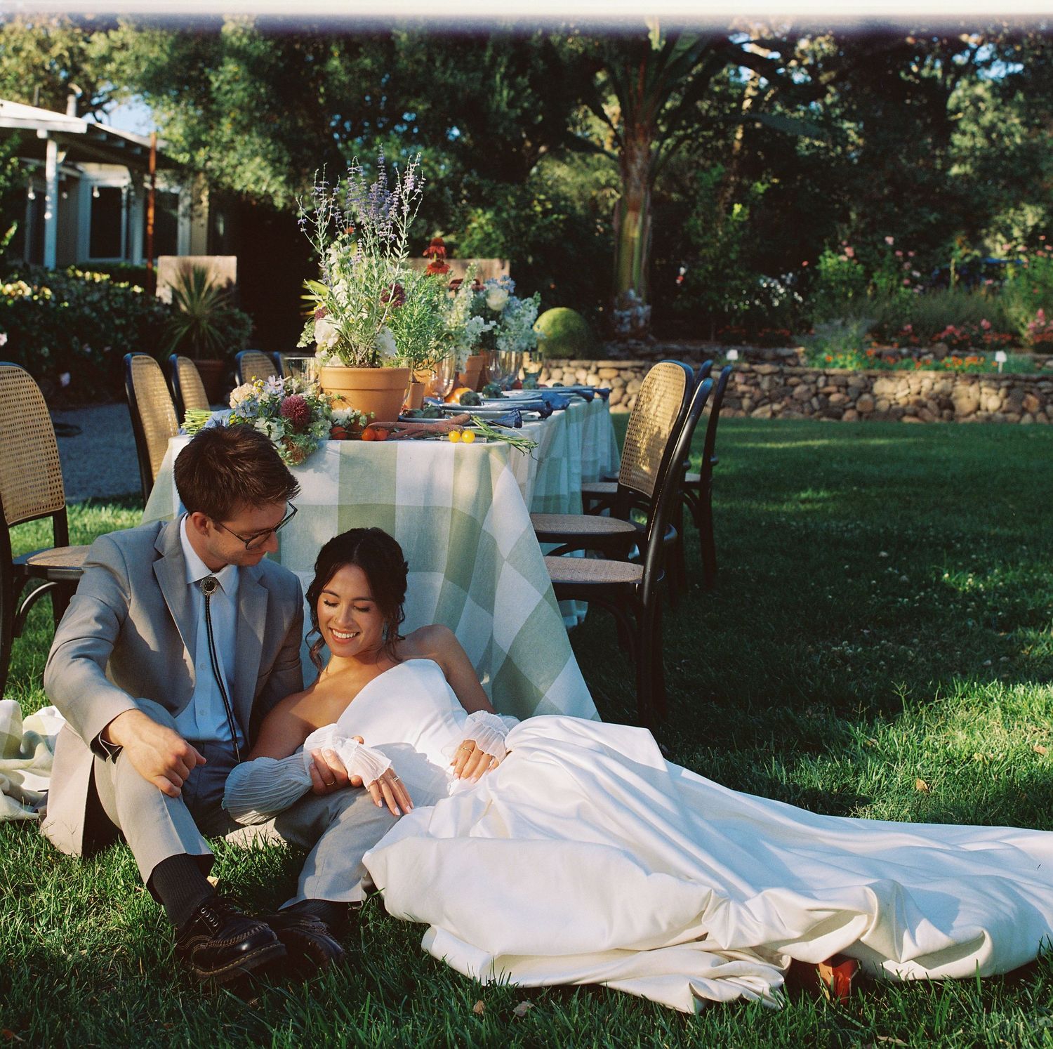 A couple in wedding attire reclines on the lawn at an outdoor garden reception with elegant table settings.