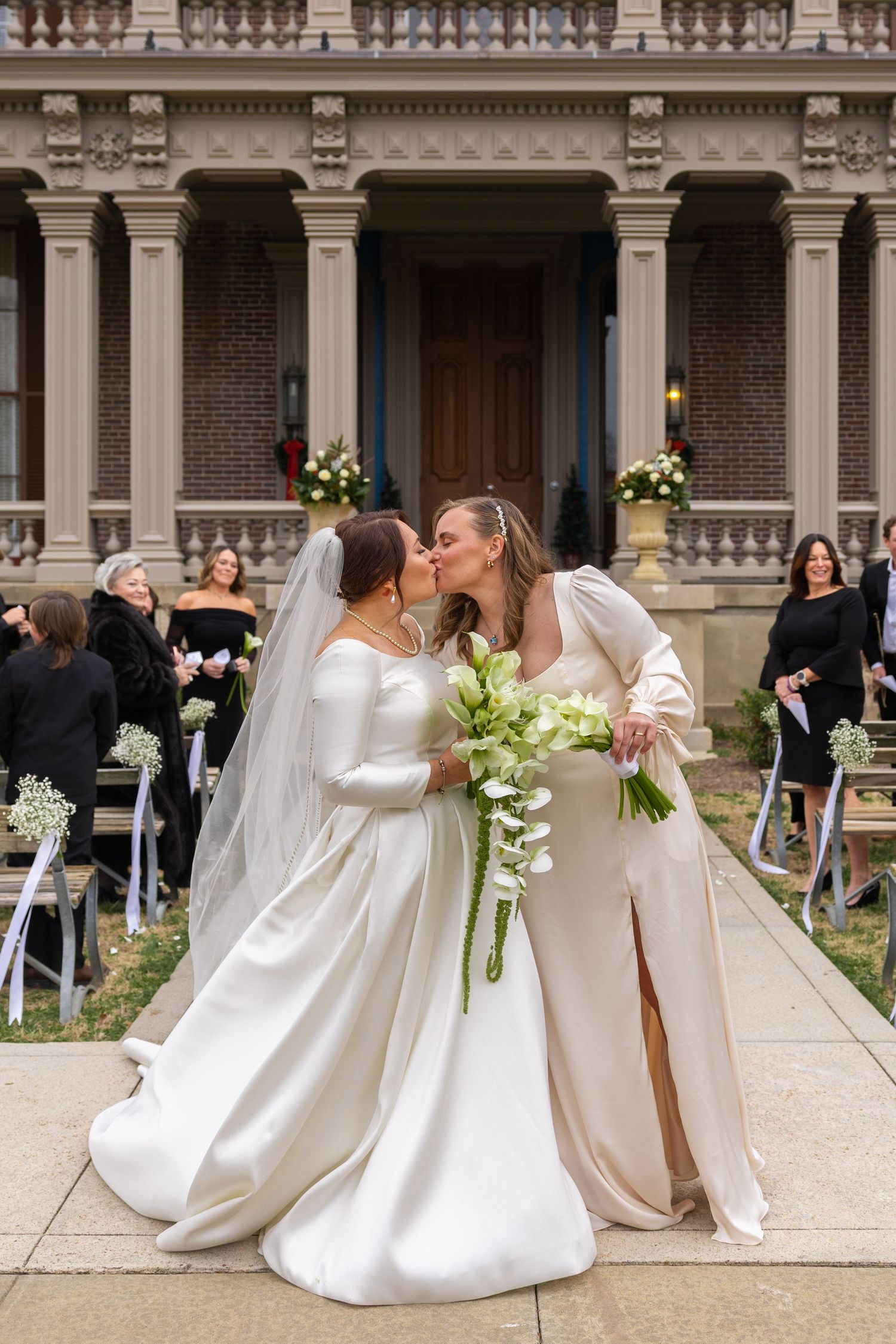 Four wedding couples in white dresses and formal attire pose together in front of a brick building during a ceremony.