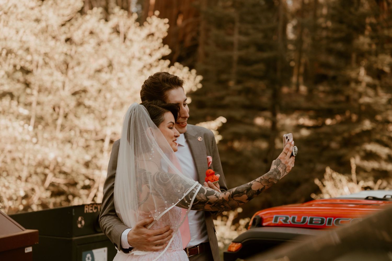 Bride and groom share a joyful moment among autumn trees with vintage red vehicle in background.