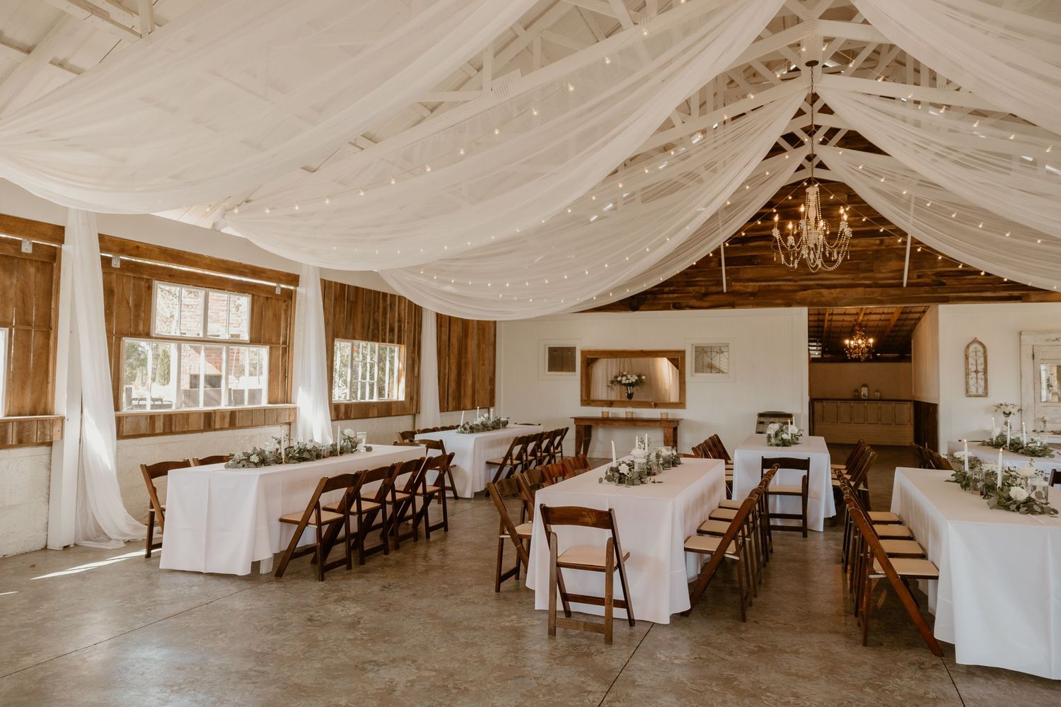Elegant wedding reception hall with white draped ceiling, chandeliers, and wooden folding chairs.