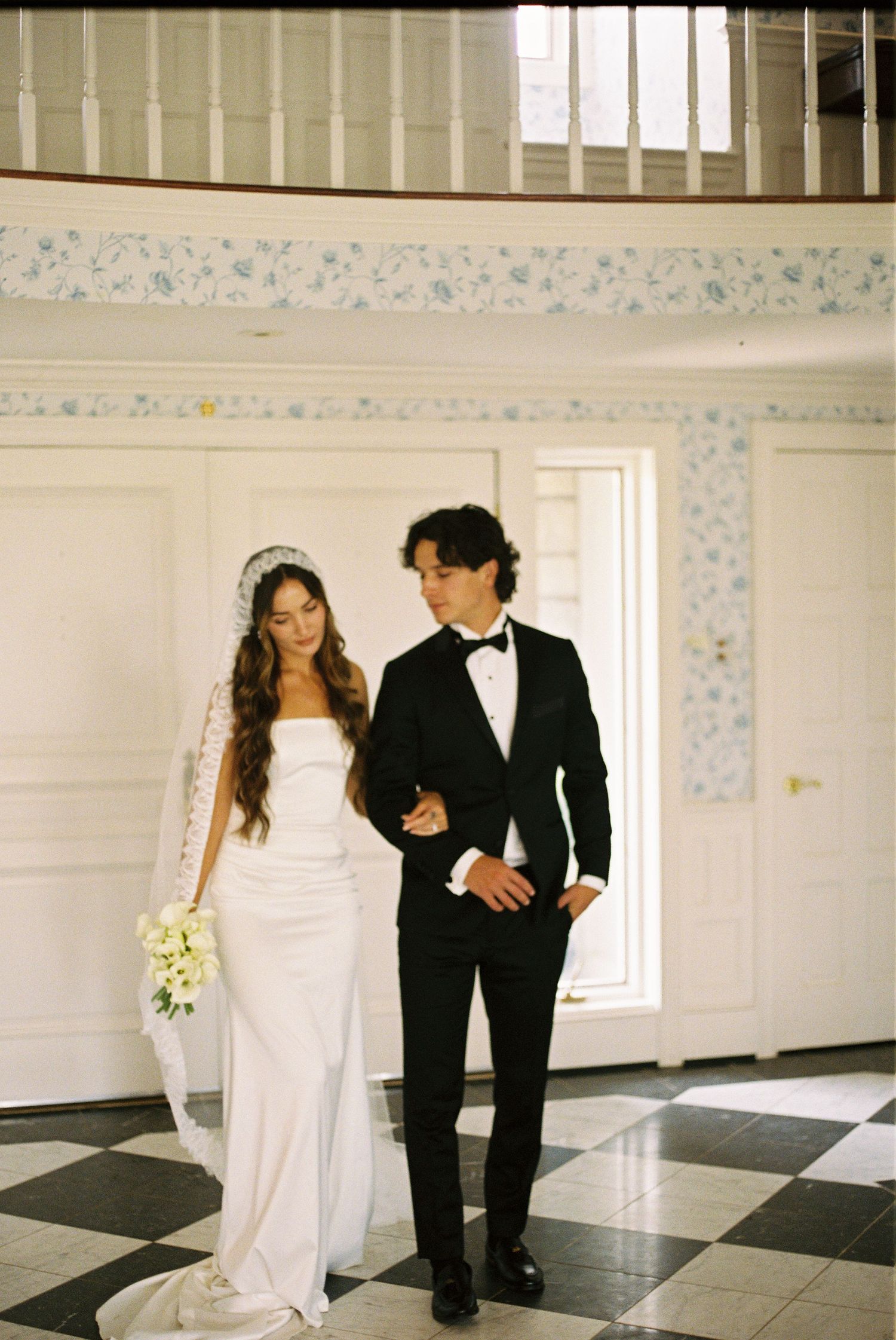 A formal couple walking together in a grand hallway with black and white checkered floors.