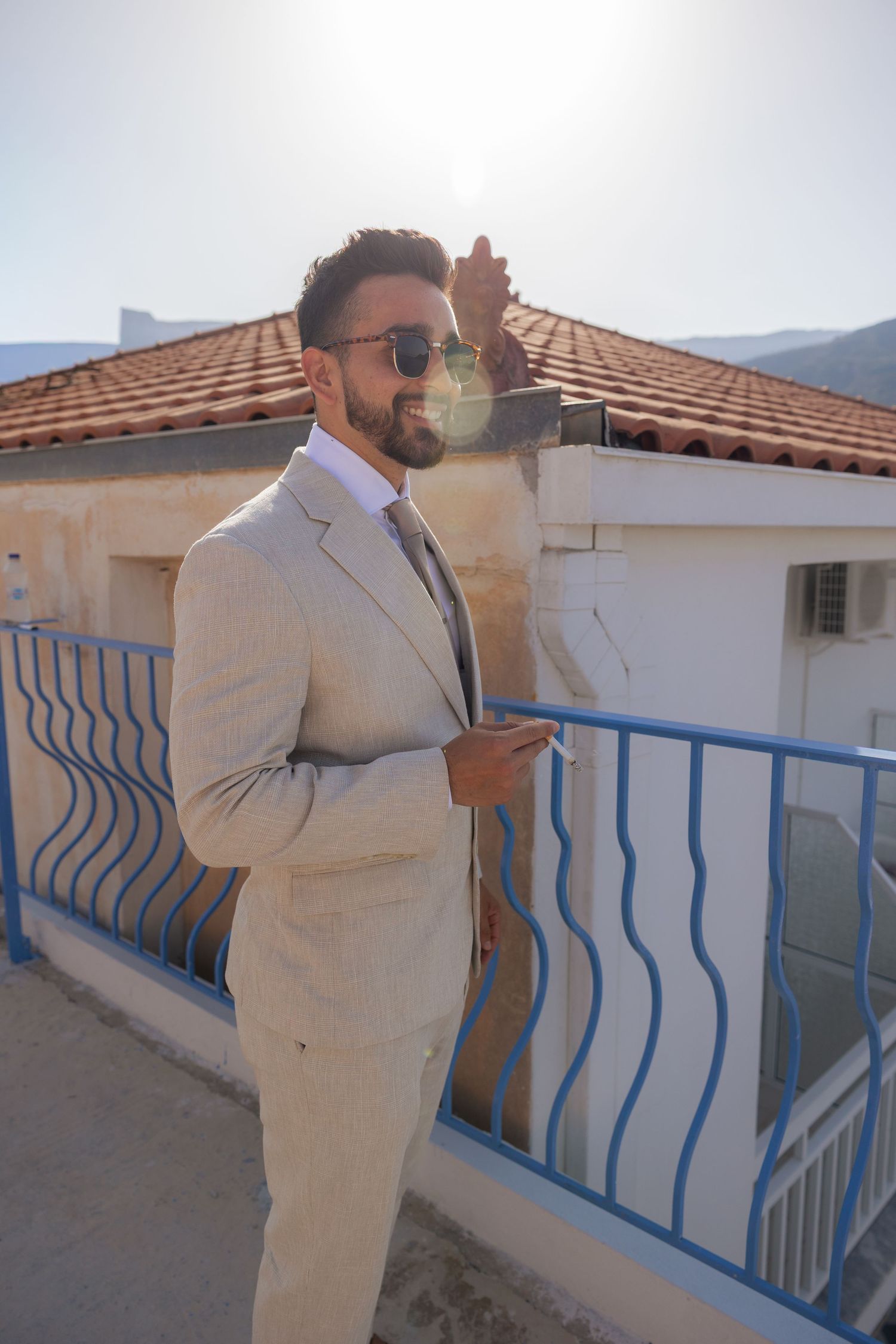 Person in a light colored suit smoking on a balcony with blue railings and terracotta roof tiles.