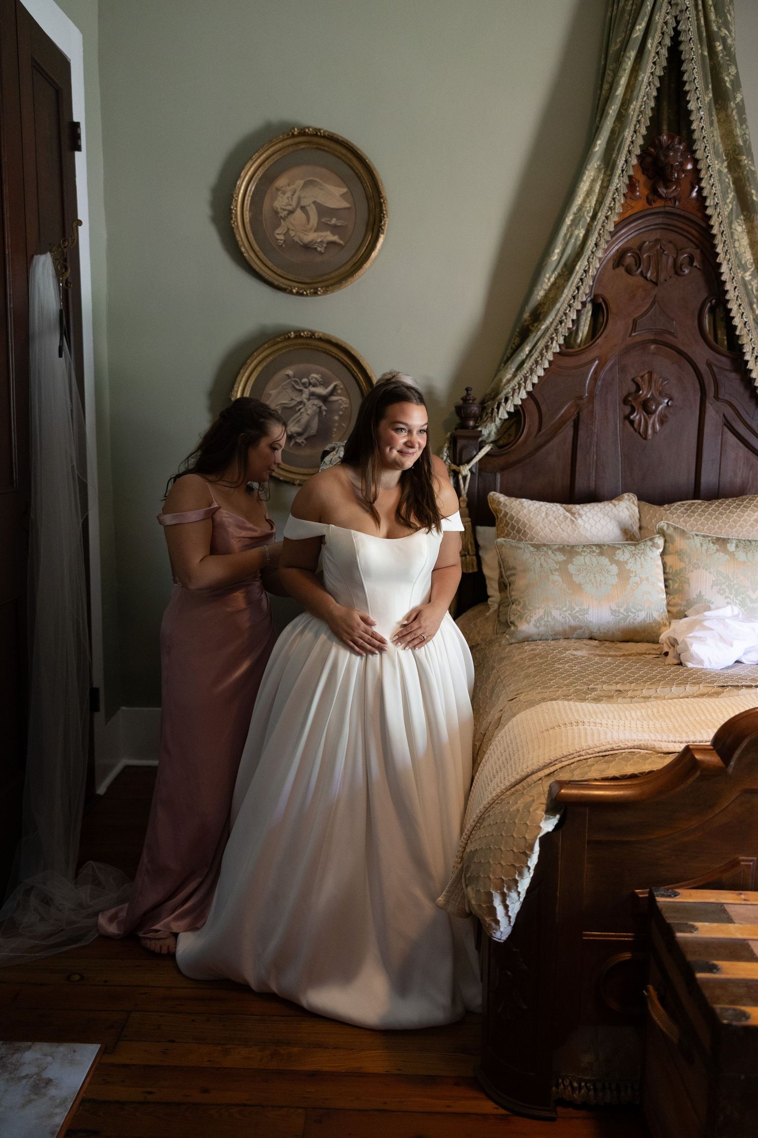 A wedding dress fitting scene in an elegant bedroom with antique decor and ornate wooden furniture.