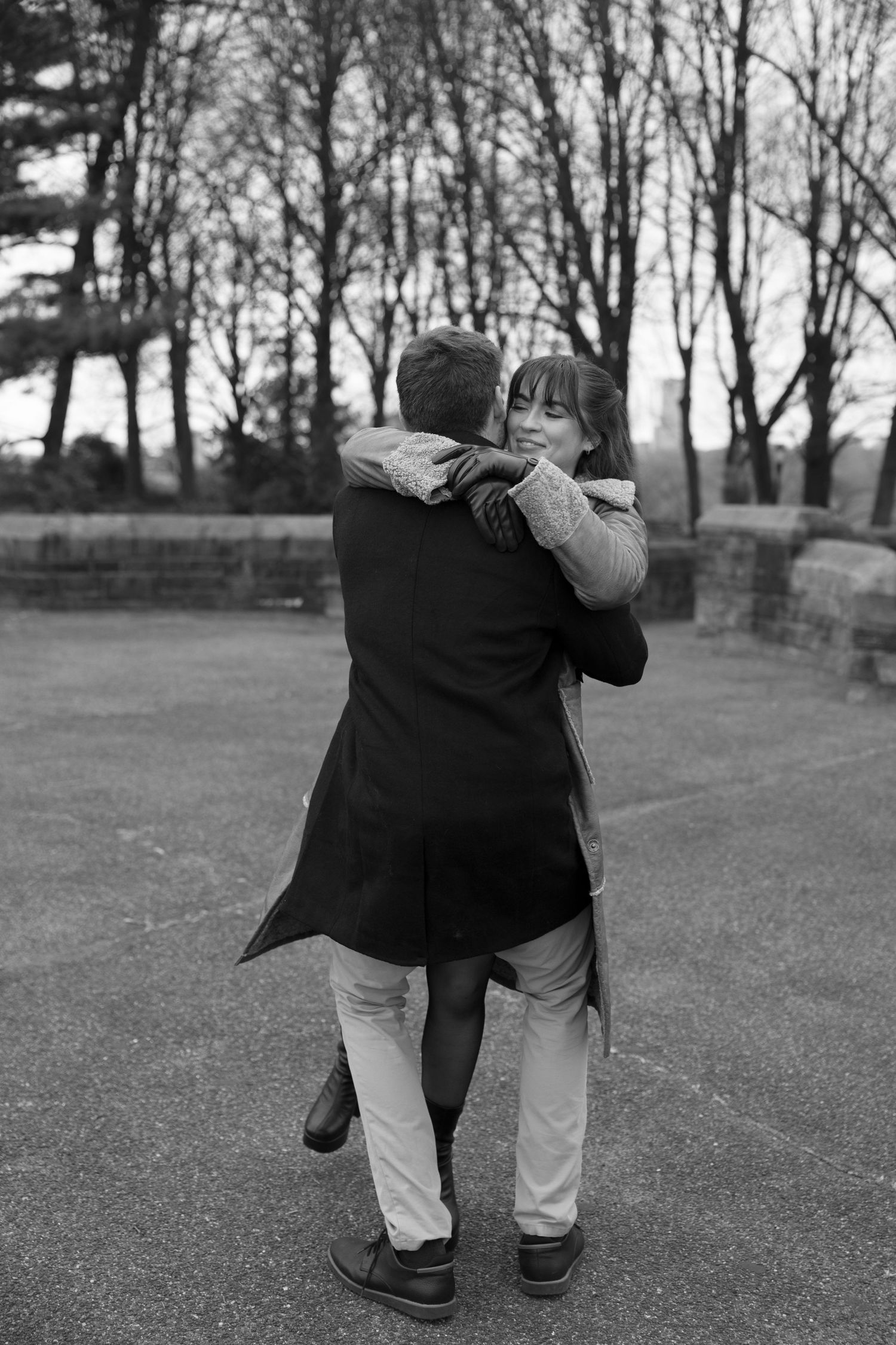 Two people in winter coats embrace while walking together on a path through bare trees in black and white.