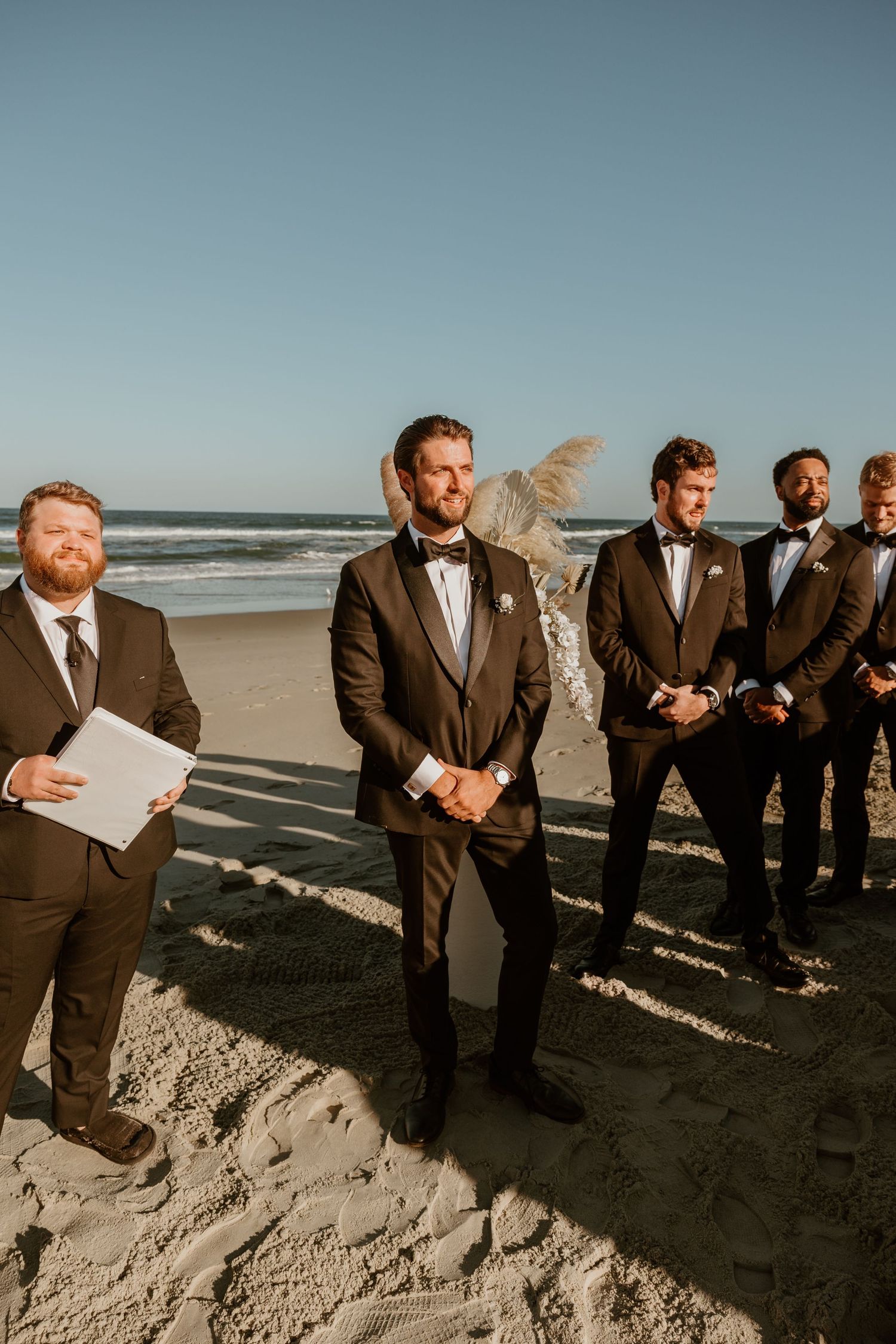Groomsmen line up on a sandy beach dune while wearing dark suits during an outdoor ceremony.