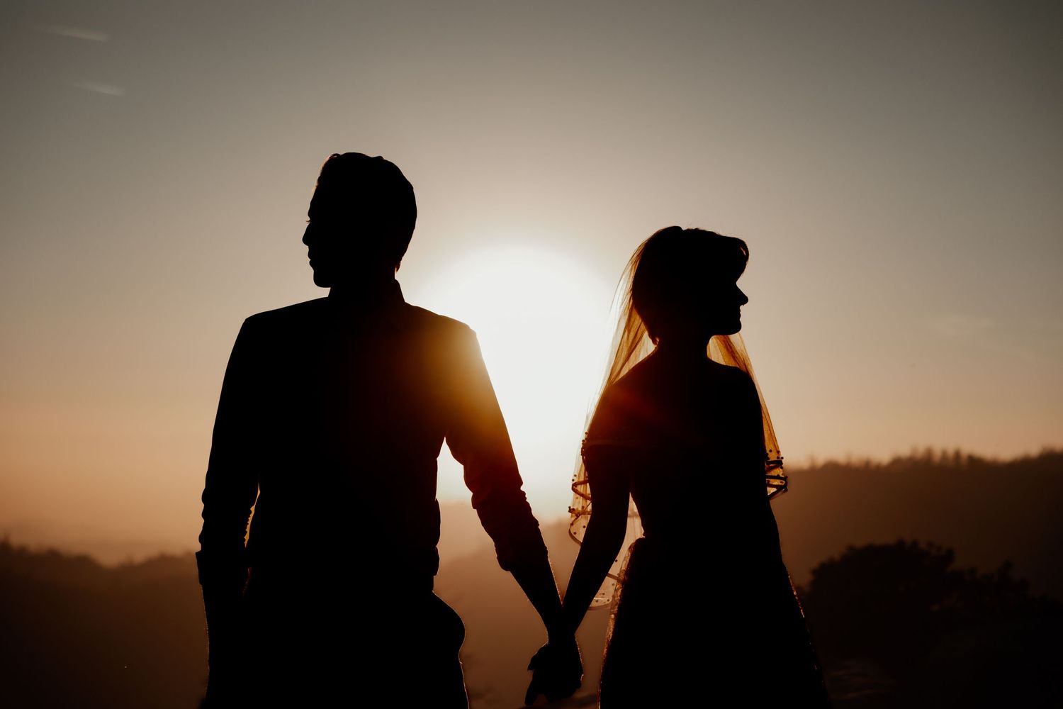 Silhouetted figures stand against dramatic sunset sky in mountain landscape.