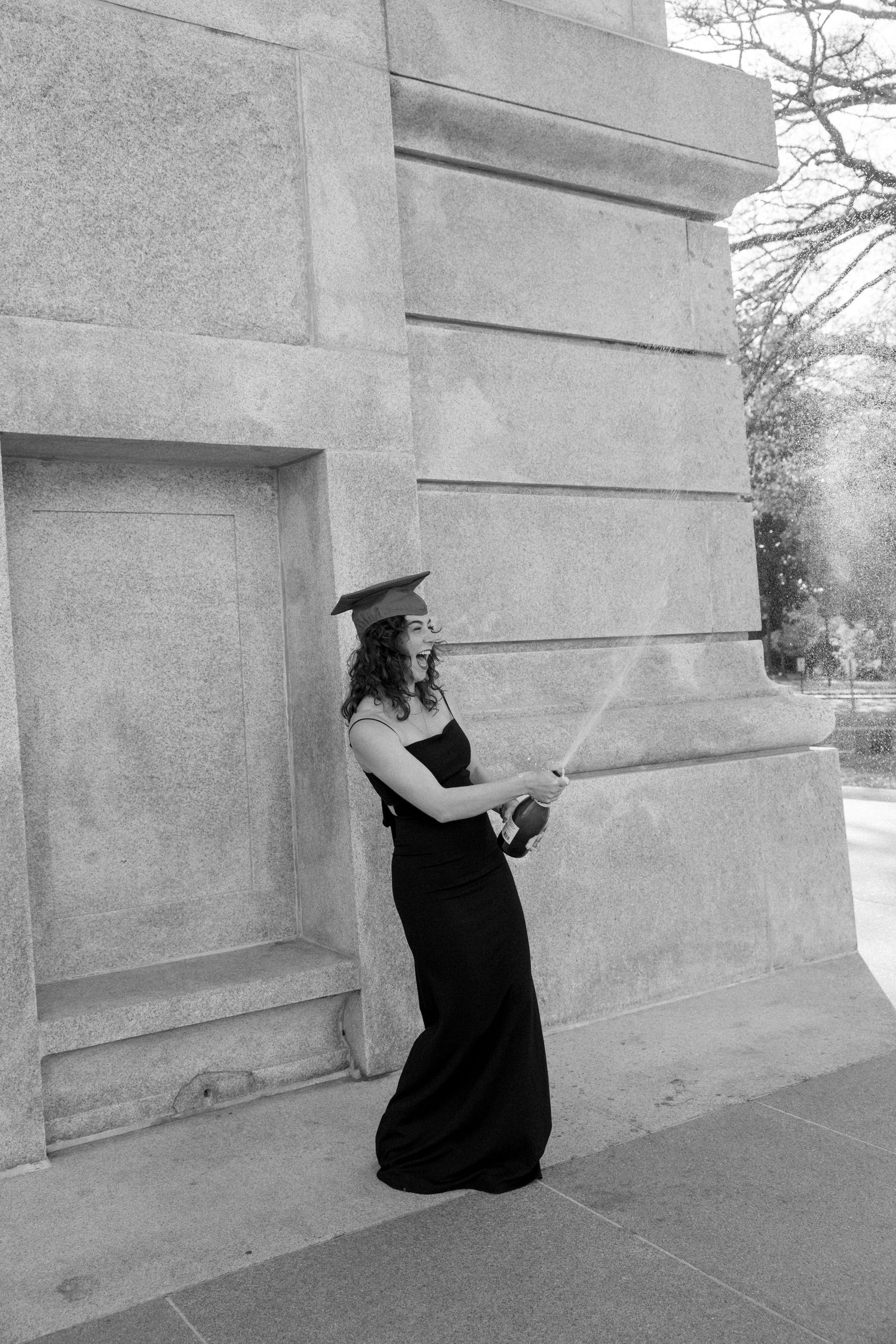 Black and white photo of a graduate in cap and gown popping champagne outside a stone building.