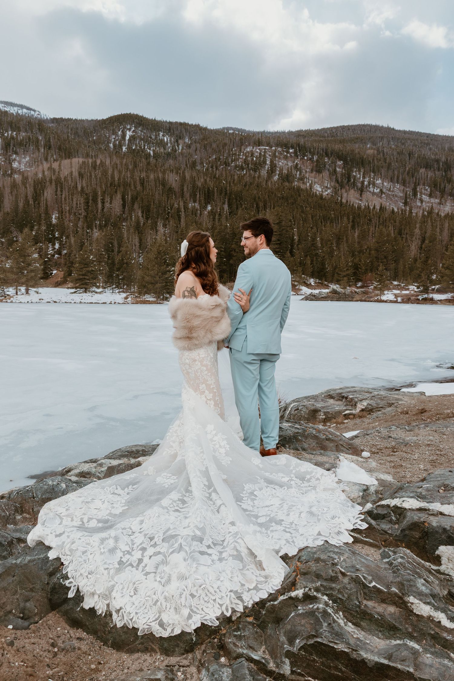 A couple in wedding attire stands by a frozen mountain lake with dramatic lace dress details.