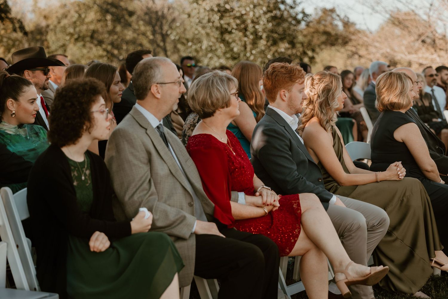 Wedding guests dressed in formal attire watch the ceremony from white folding chairs.