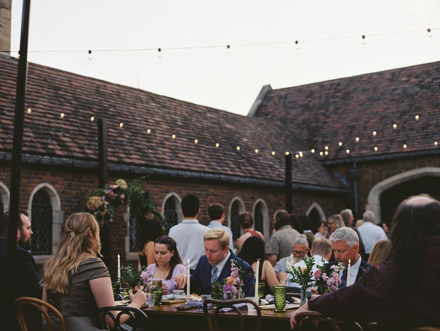 Outdoor wedding celebration with string lights illuminating guests seated at long tables under dusk sky.