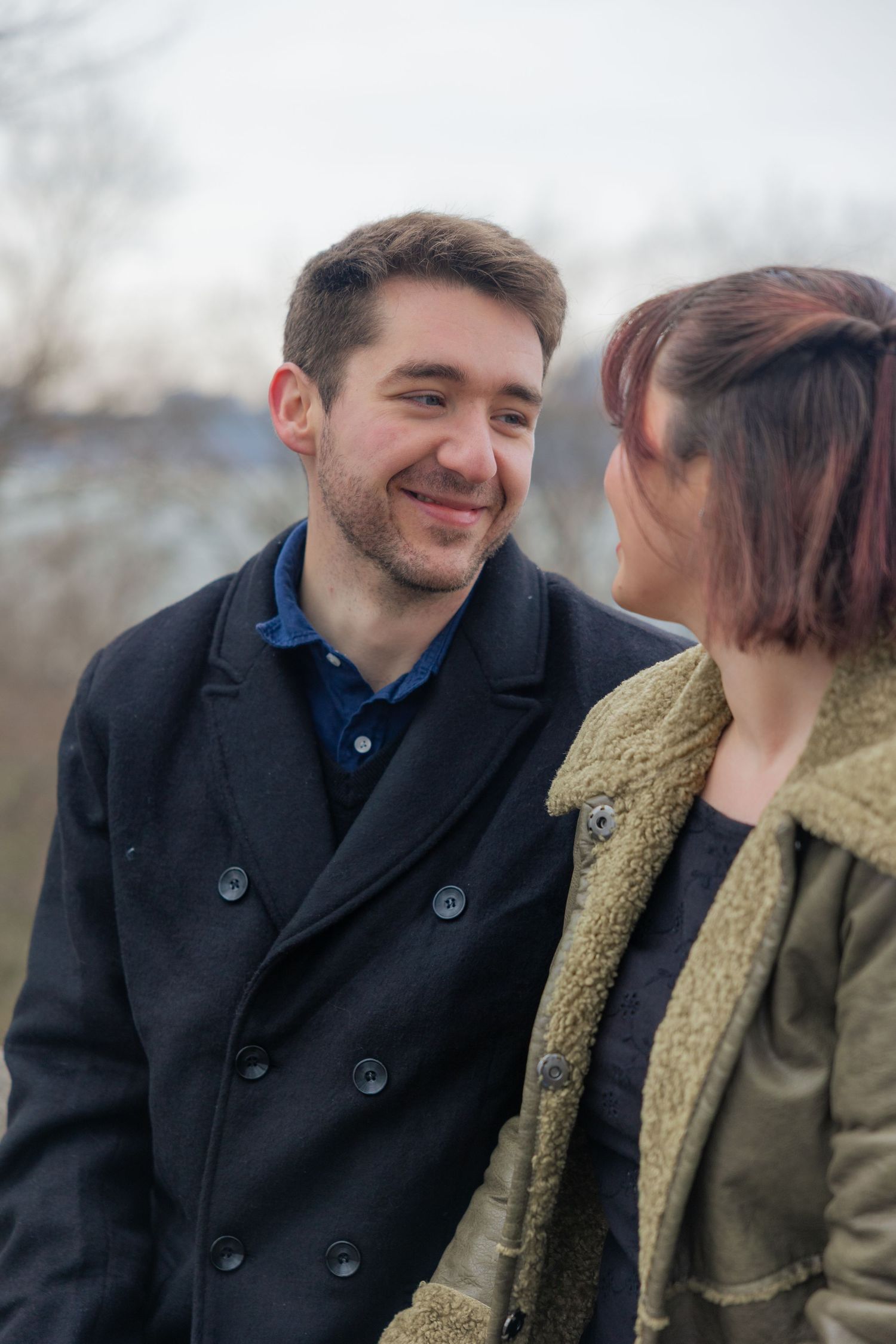 A couple shares a tender moment outdoors while wearing winter coats on an overcast day.