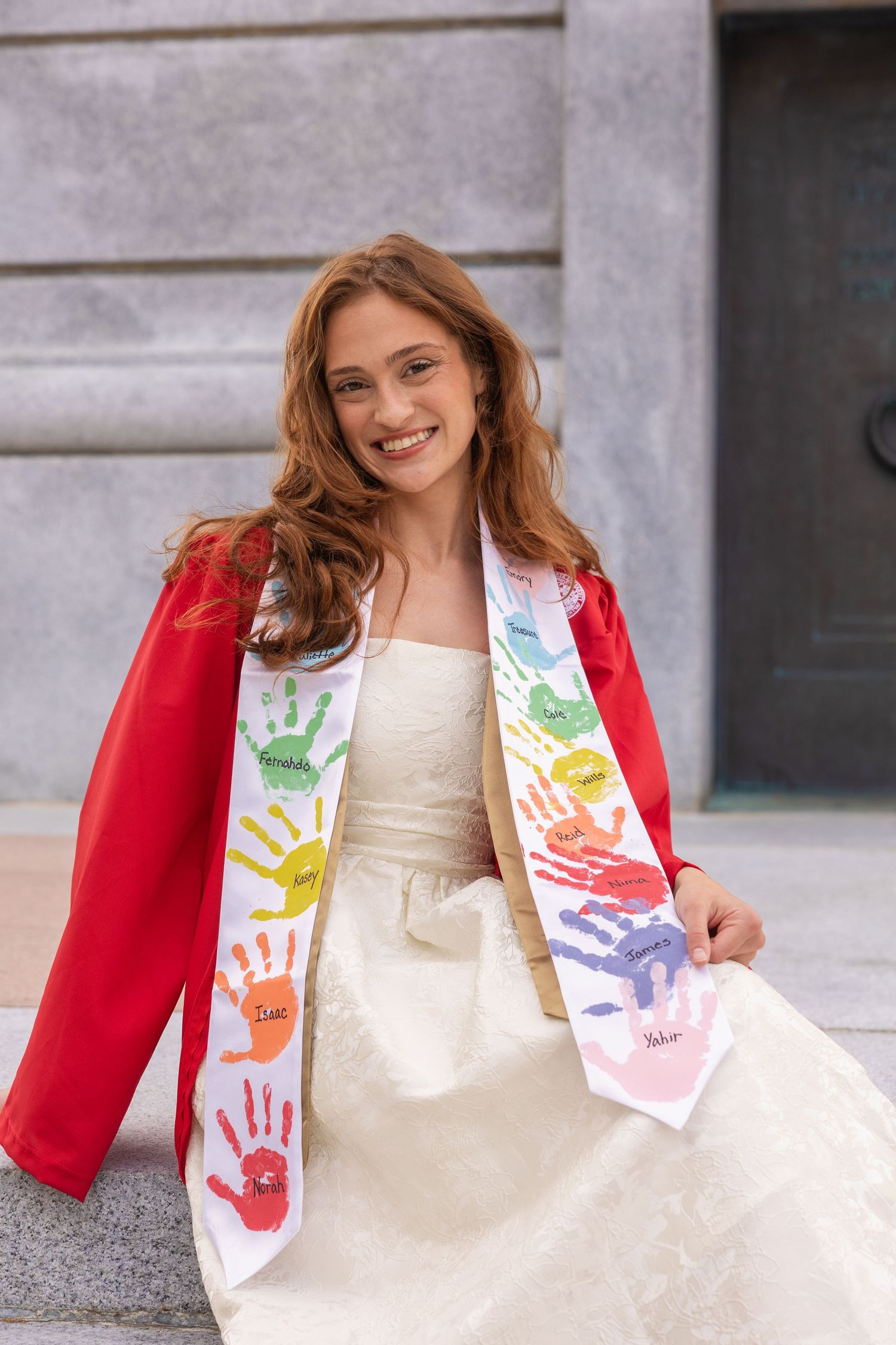 Smiling graduate in red gown wearing colorful handprint stole, sitting outdoors in white dress.