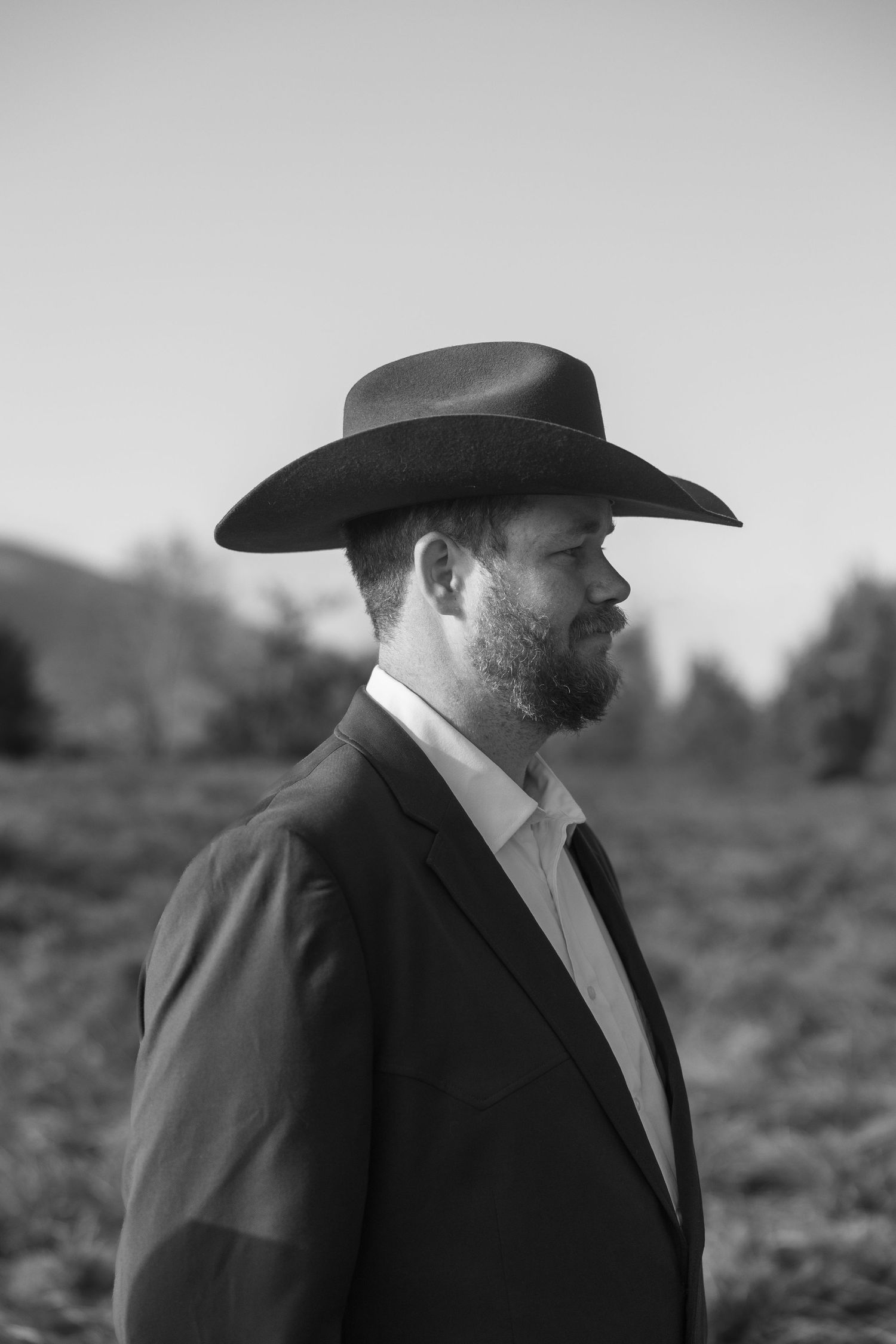 Black and white portrait of a person in a cowboy hat and suit jacket looking to the side against a blurred background.