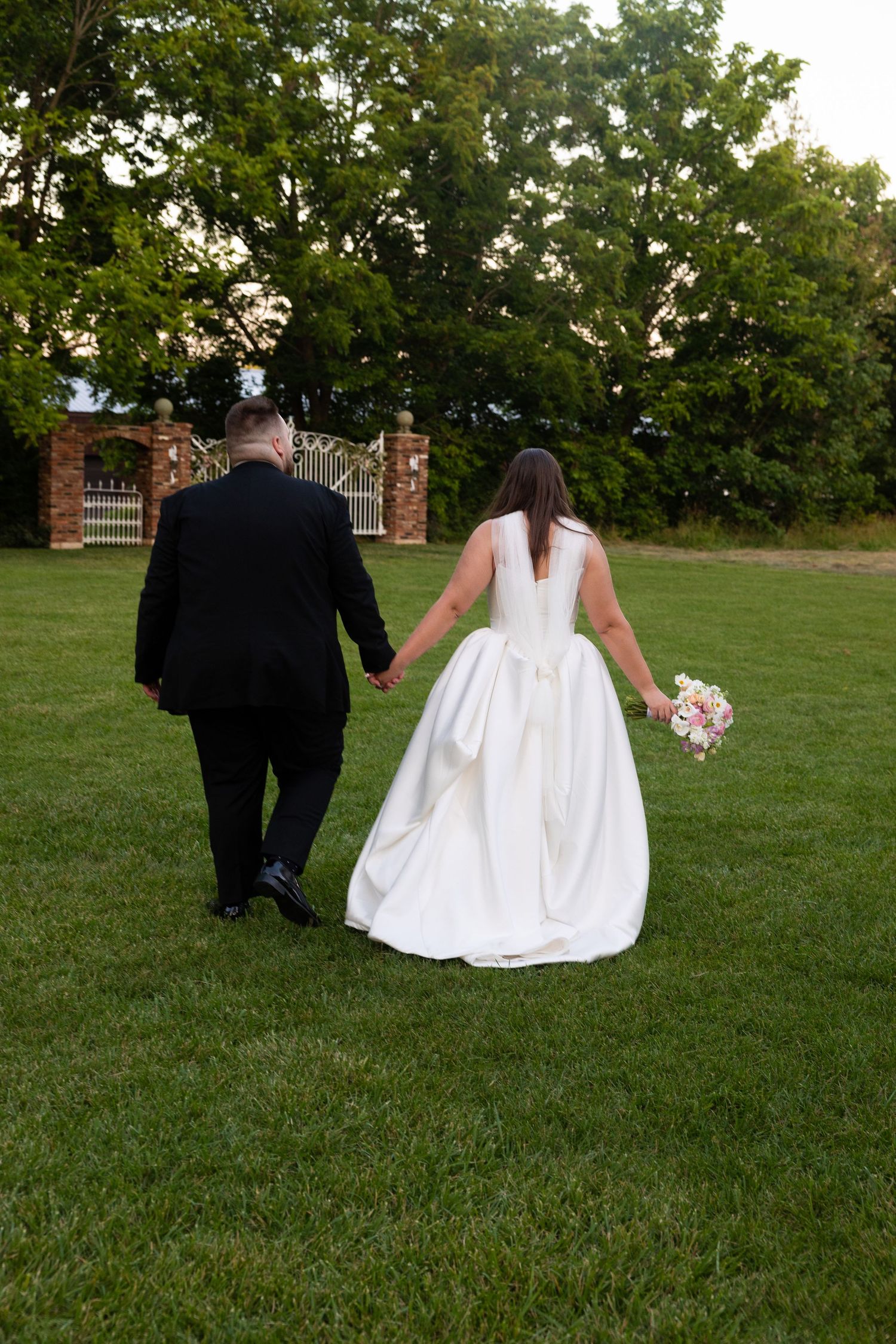 Newlyweds walk hand in hand across a lush green lawn with brick pillars in the background.