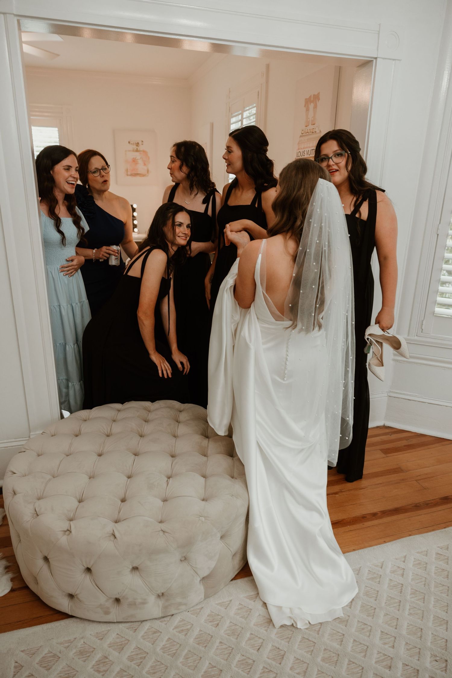 Wedding party gathering around a white tufted ottoman in an elegant room.
