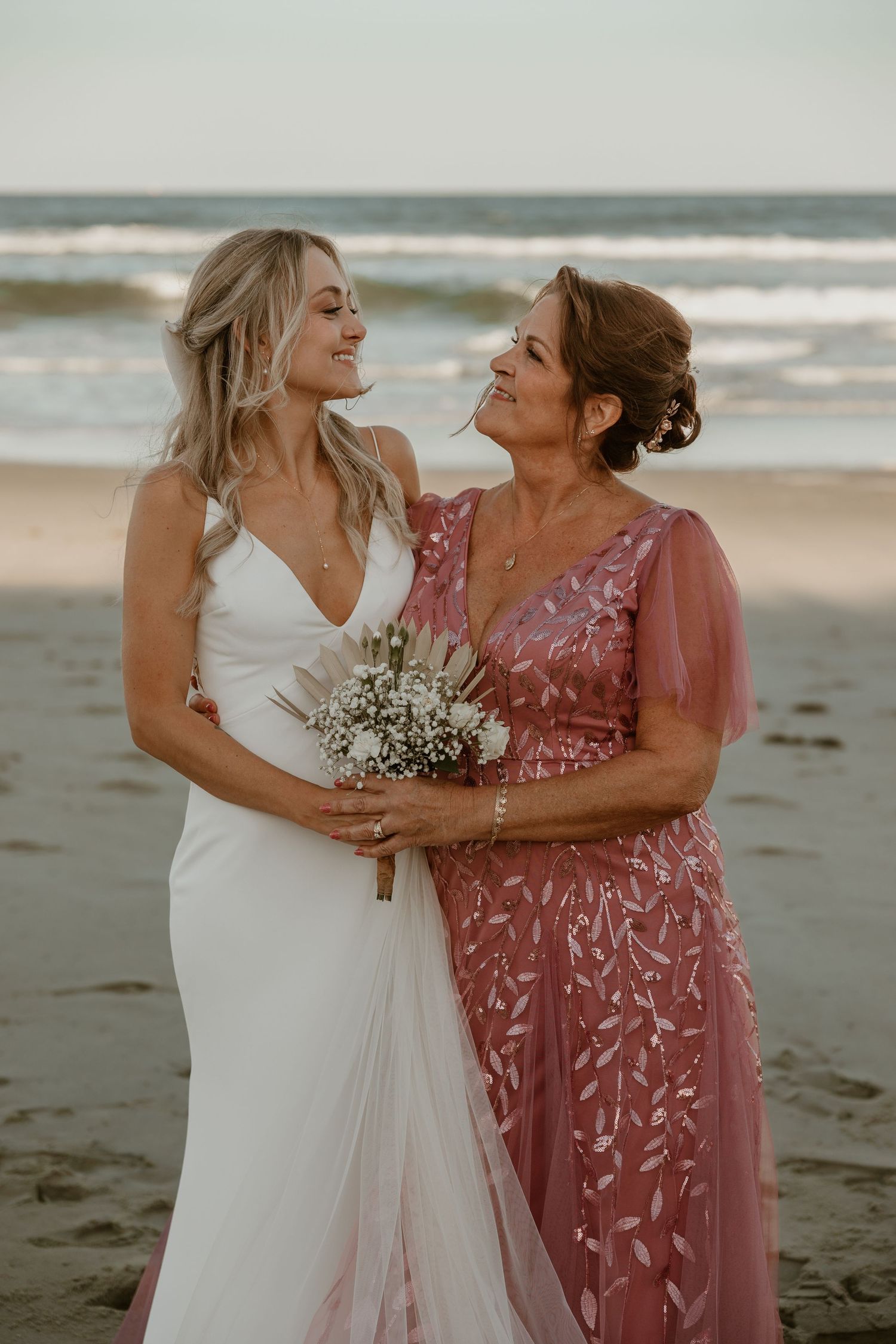 Tender moment between bride and woman in pink dress sharing bouquet on beach.