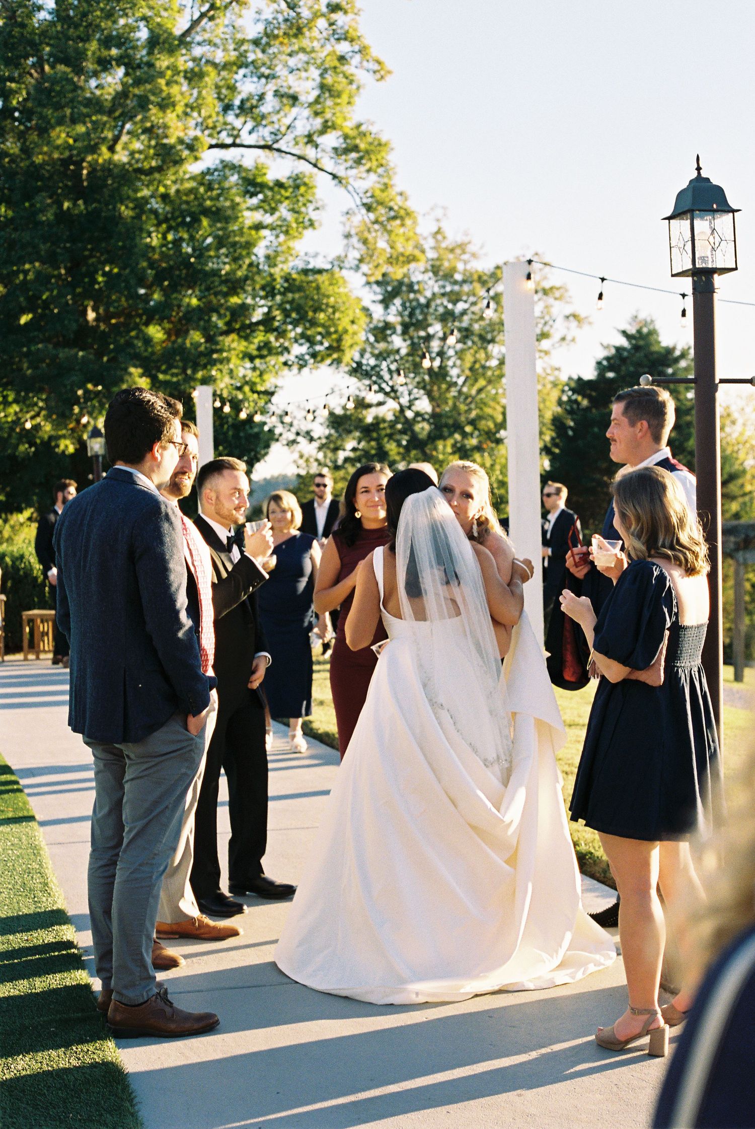 Wedding party gathers outdoors on a paved walkway during golden hour.