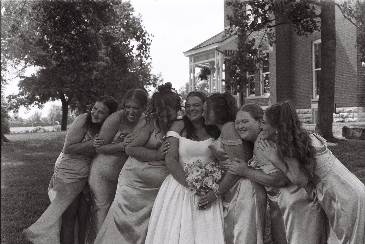 A group of bridesmaids in light-colored dresses pose together outdoors near a brick building.