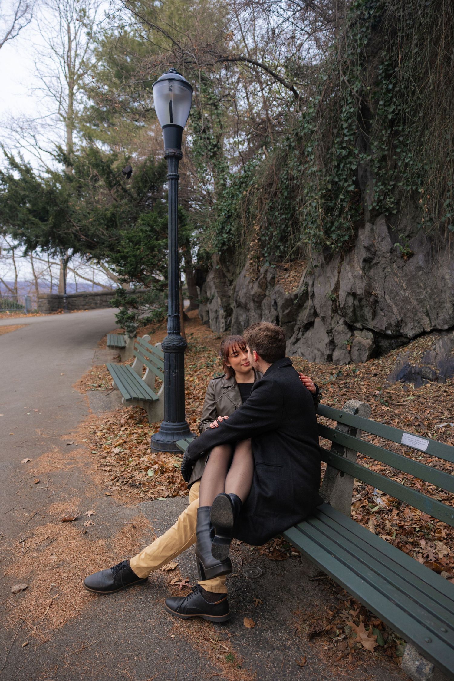 Person sitting alone on a park bench next to a vintage lamp post on an autumn path.