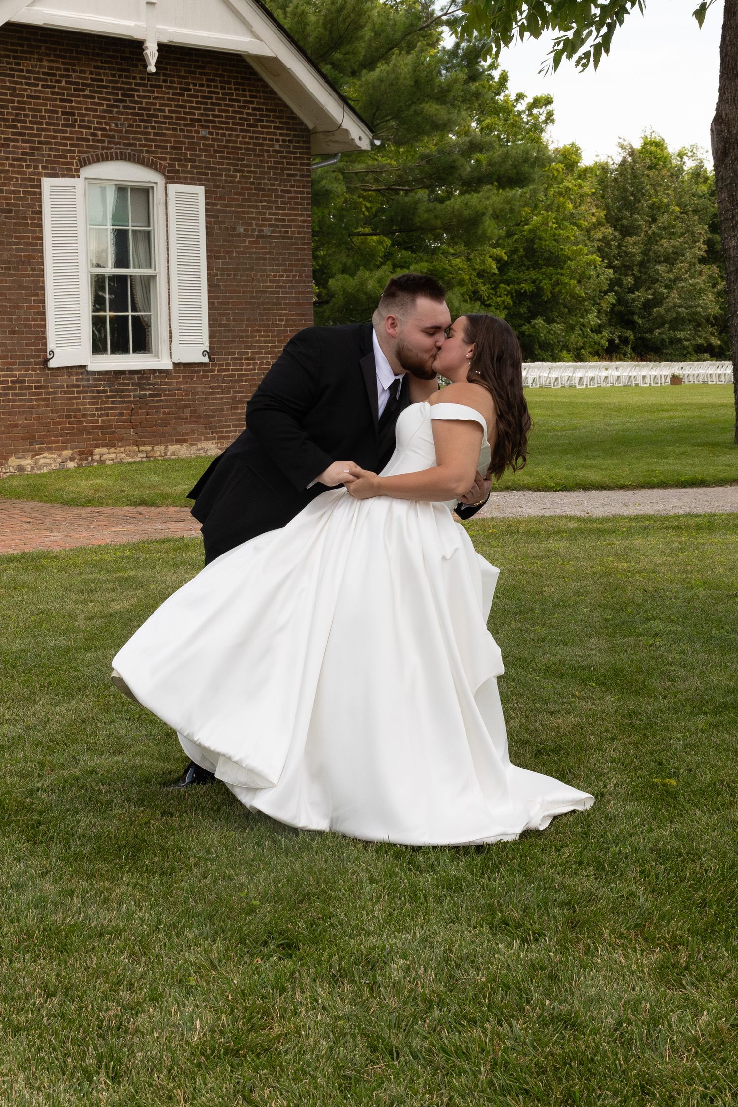 Romantic wedding couple embracing on lawn in front of brick building with white shutters.