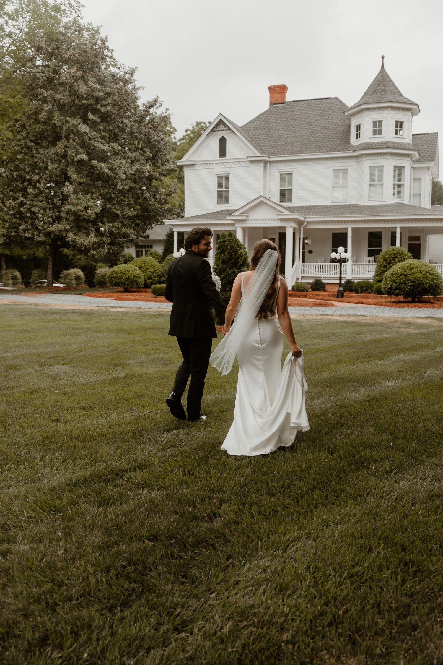 A wedding couple walks on the lawn of a classic white Victorian-style house.