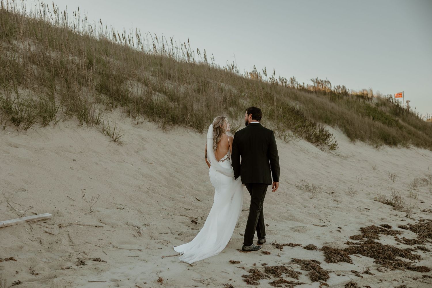 A couple in wedding attire walks up a sandy dune path together.