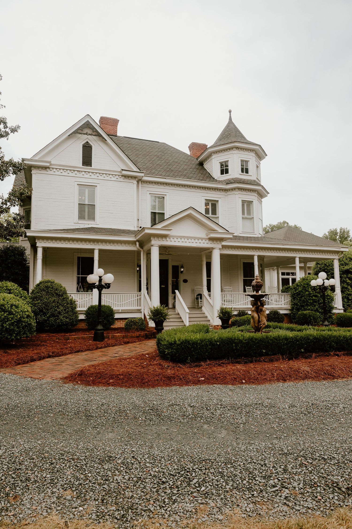 A classic white Victorian-style house with wraparound porch and gravel driveway.