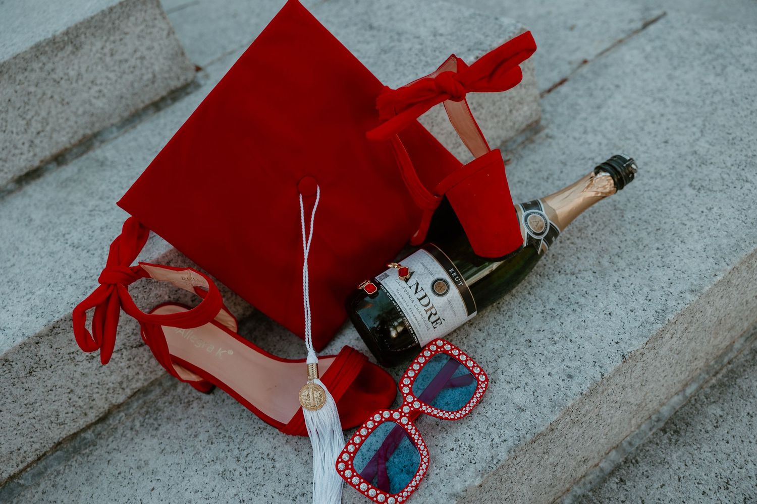 Red gift bag with champagne bottle and heart-shaped sunglasses on stone steps, perfect for a romantic celebration.