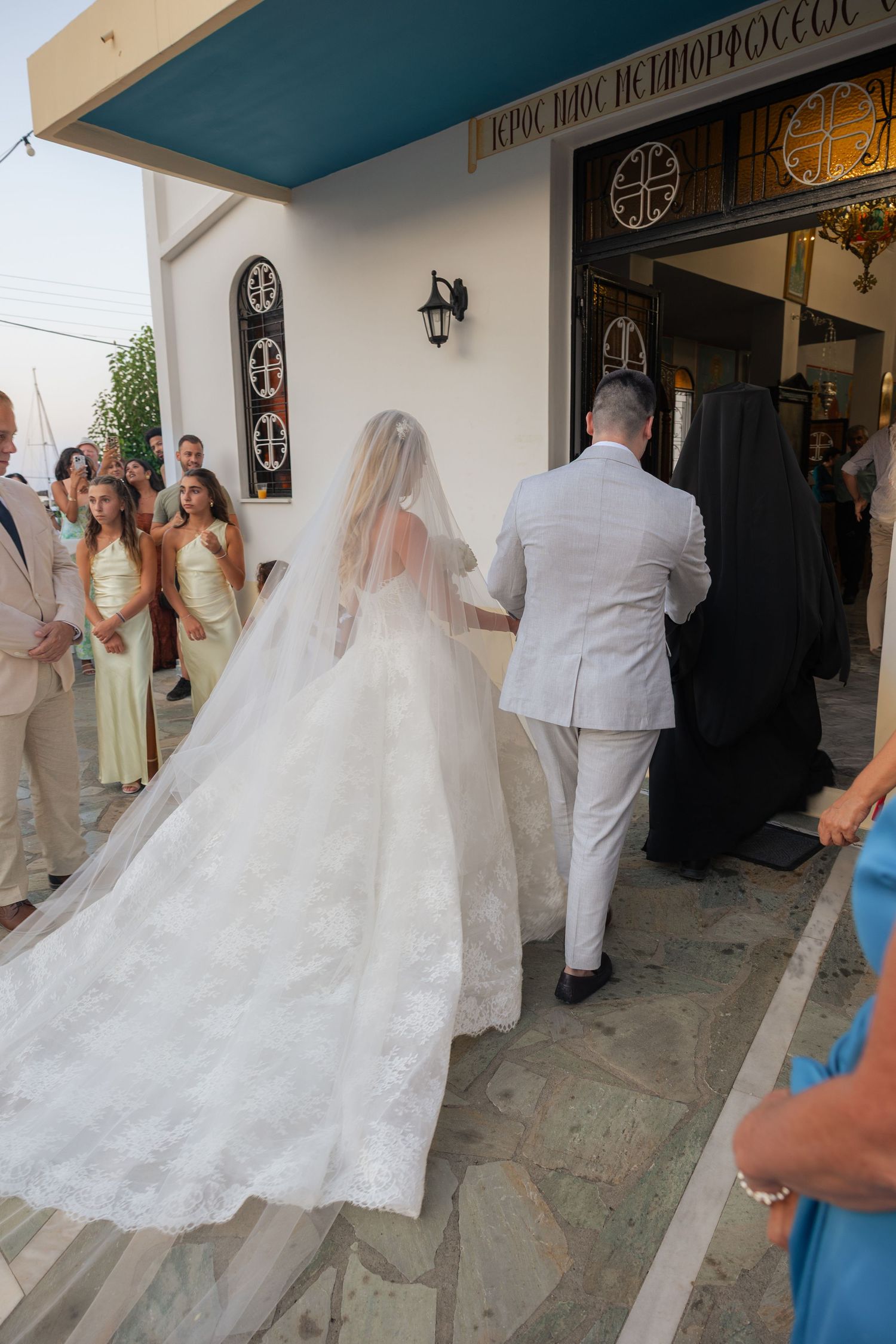 A bride in a flowing white lace gown with a long train enters a Greek Orthodox church for her wedding ceremony.