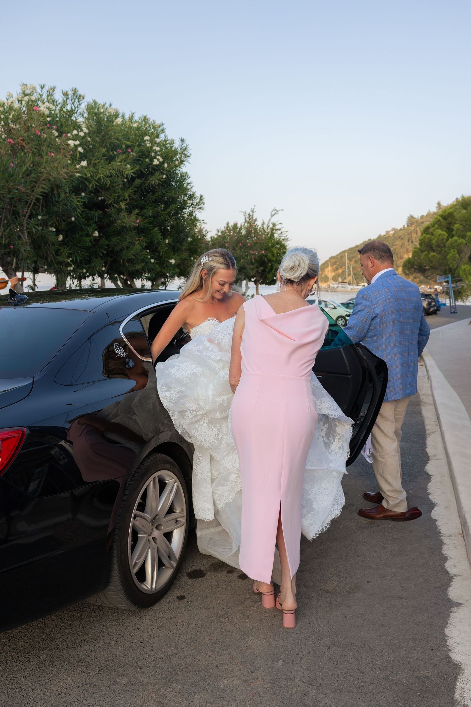 A group of people in formal attire lean into a black car parked on a road with trees in the background.
