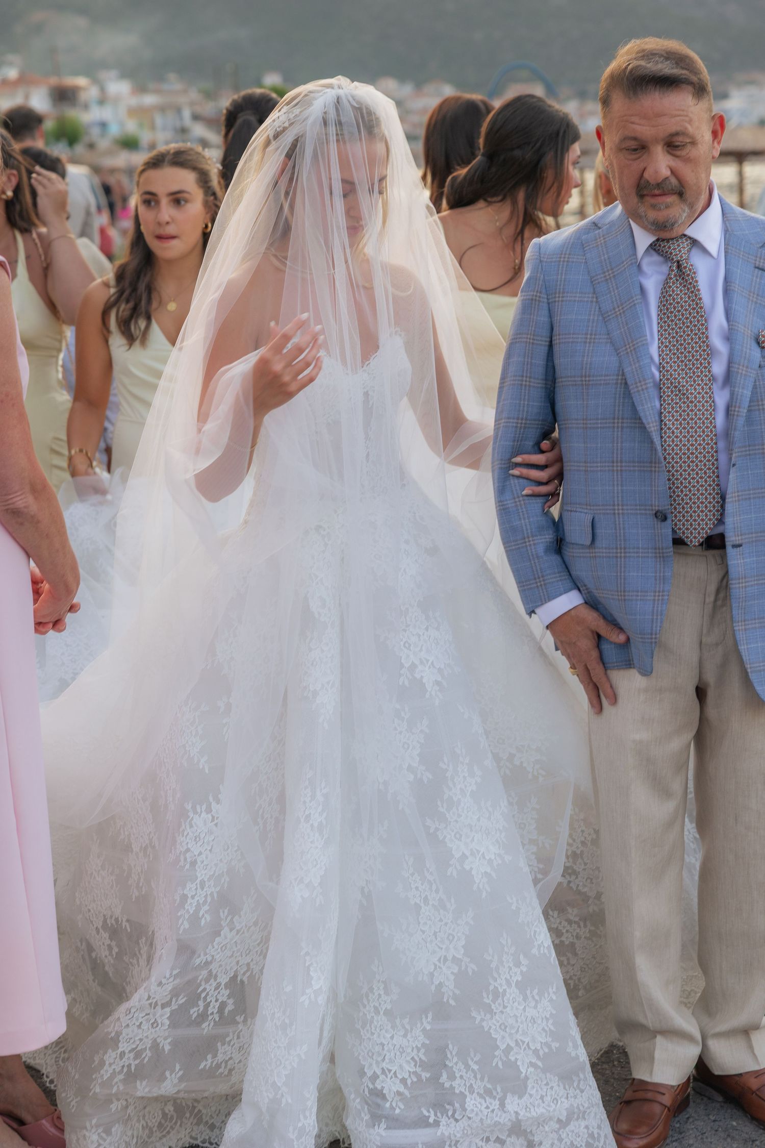 A bride in a flowing lace wedding dress with a long veil walks alongside a man in a light blue checkered suit.