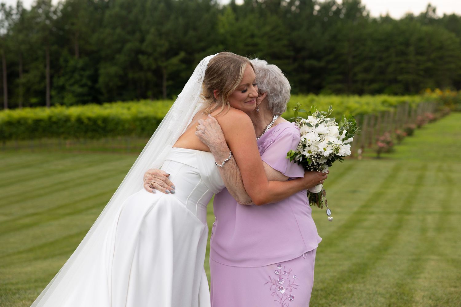 A bride in white dress embraces someone in a pink dress while holding a bouquet outdoors on manicured lawn.