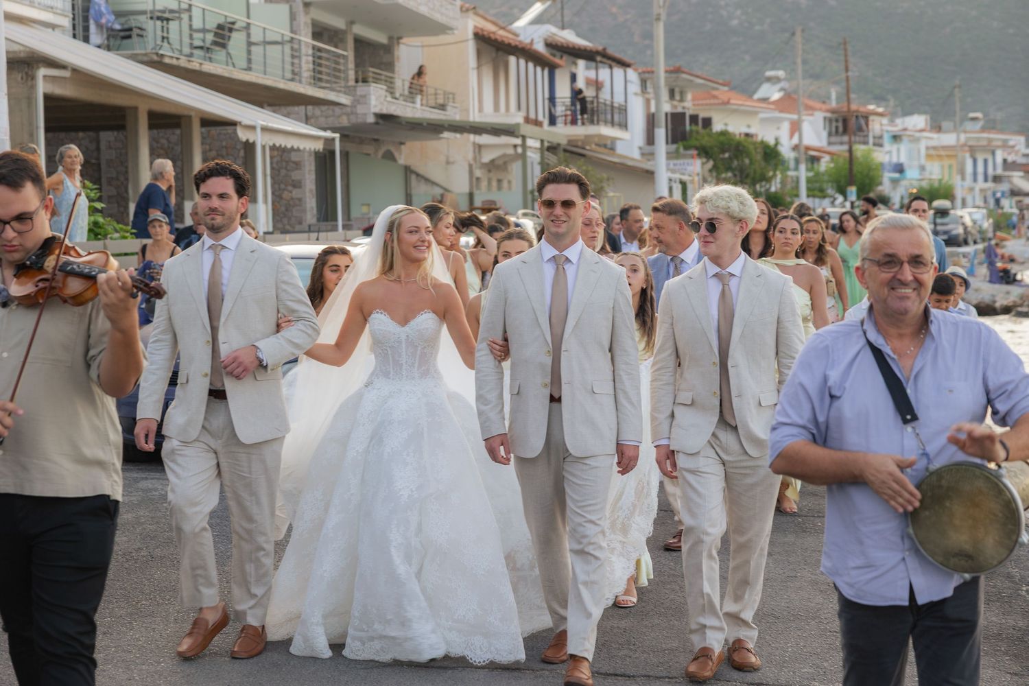 A wedding party procession walks down a street in Greece with musicians and guests dressed in white and beige attire.