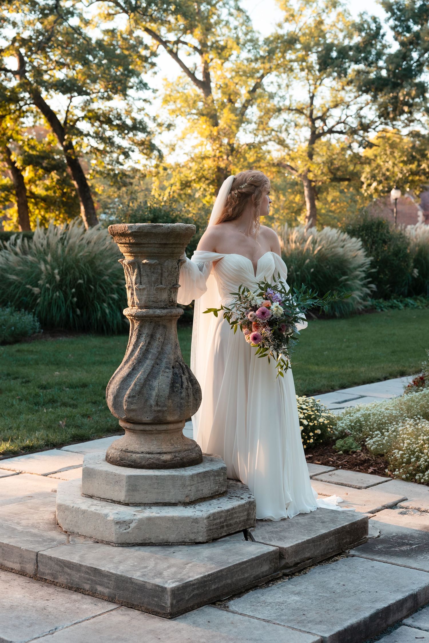 Wedding dress and bouquet photographed against stone urns and garden setting at sunset.