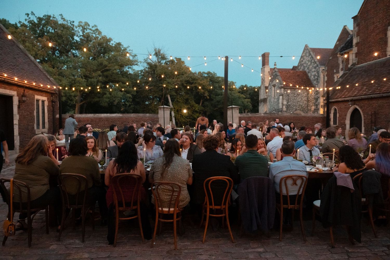 Outdoor wedding reception at dusk with string lights illuminating tables of guests under a warm evening sky.