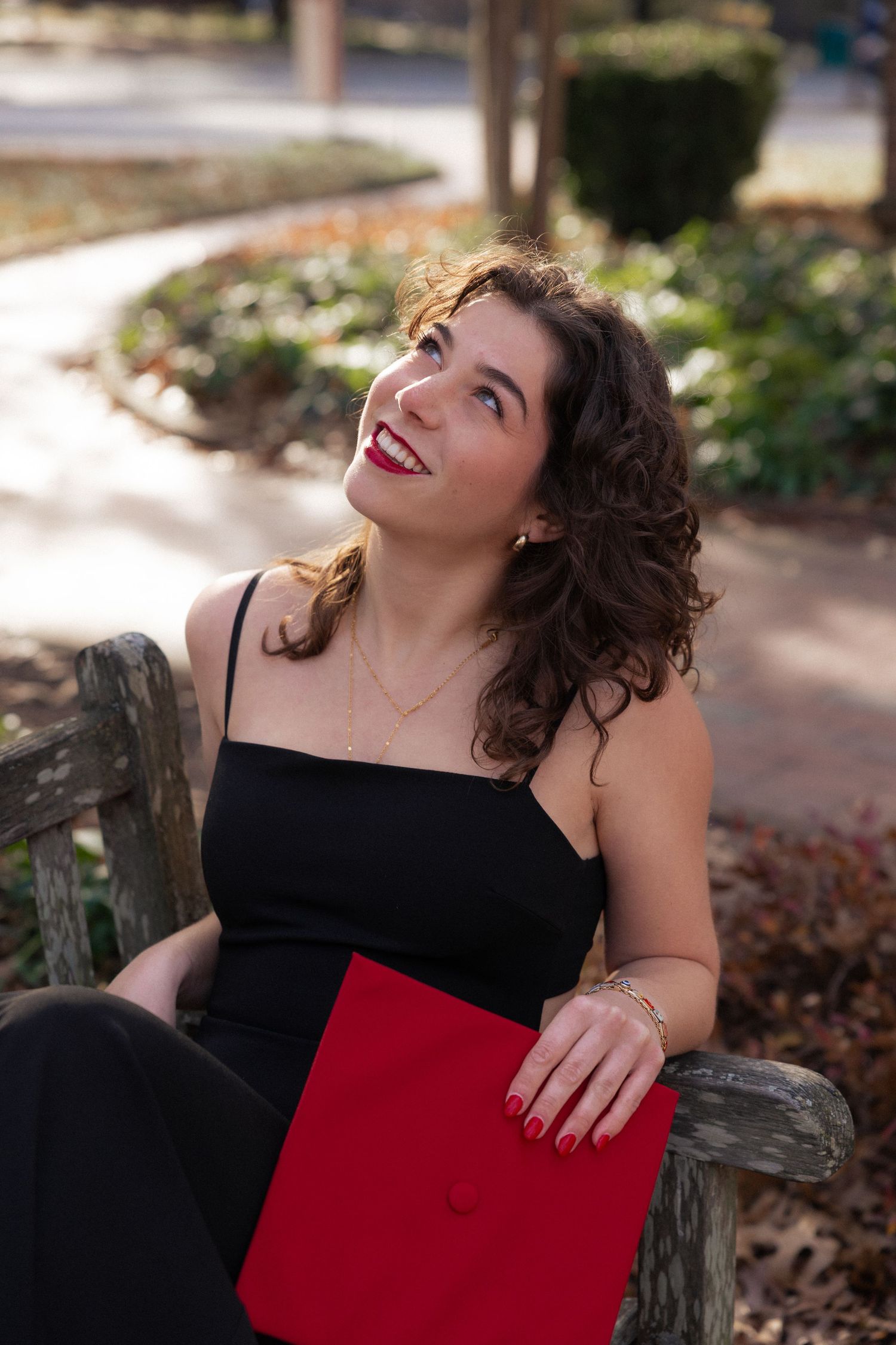 Young woman in black dress holding red diploma folder, smiling on park bench during graduation photo session.