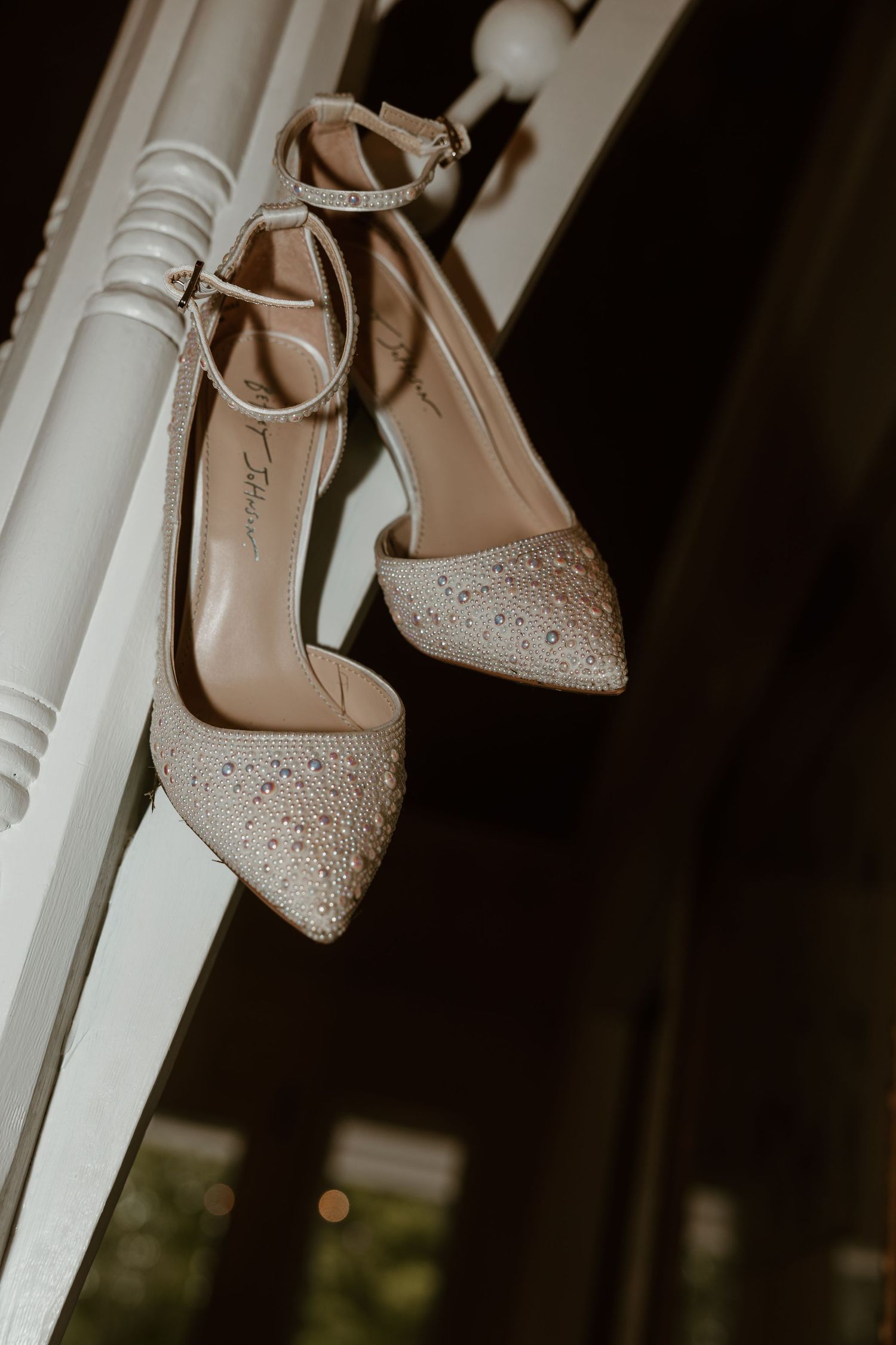 Elegant beige wedding shoes with rhinestone details hanging on a white railing.