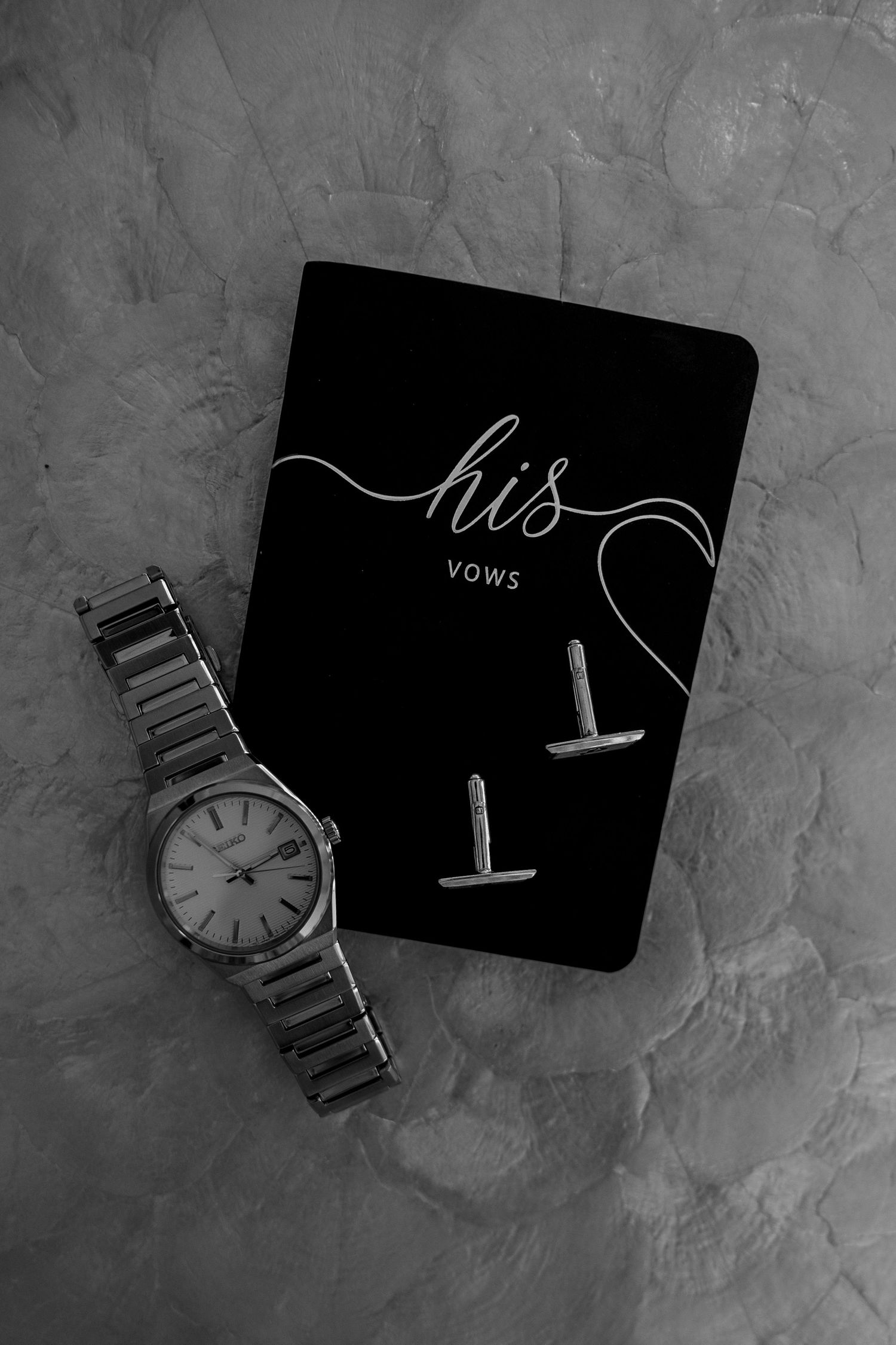 Black vow book with 'his' text and silver watch on textured gray surface.