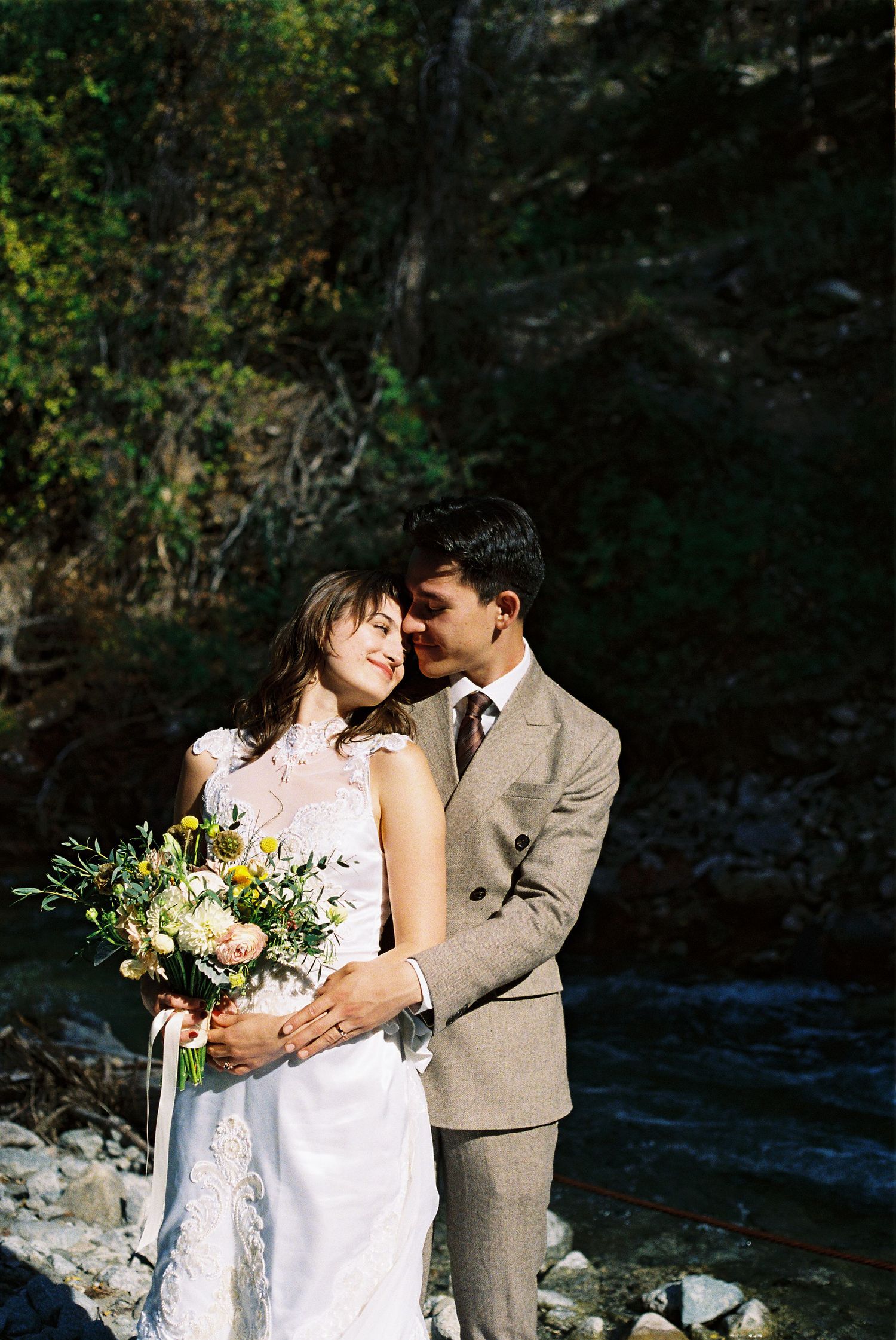 An intimate moment between newlyweds by a stream, with the bride holding a pastel bouquet.