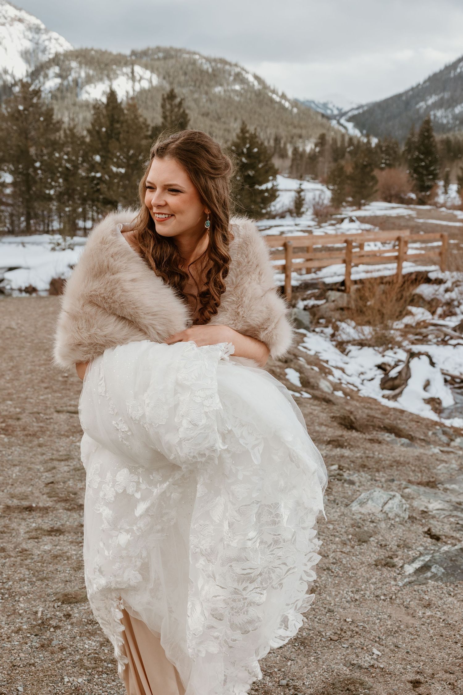 A bride in a lace dress and fur wrap spins in front of a snowy mountain landscape.
