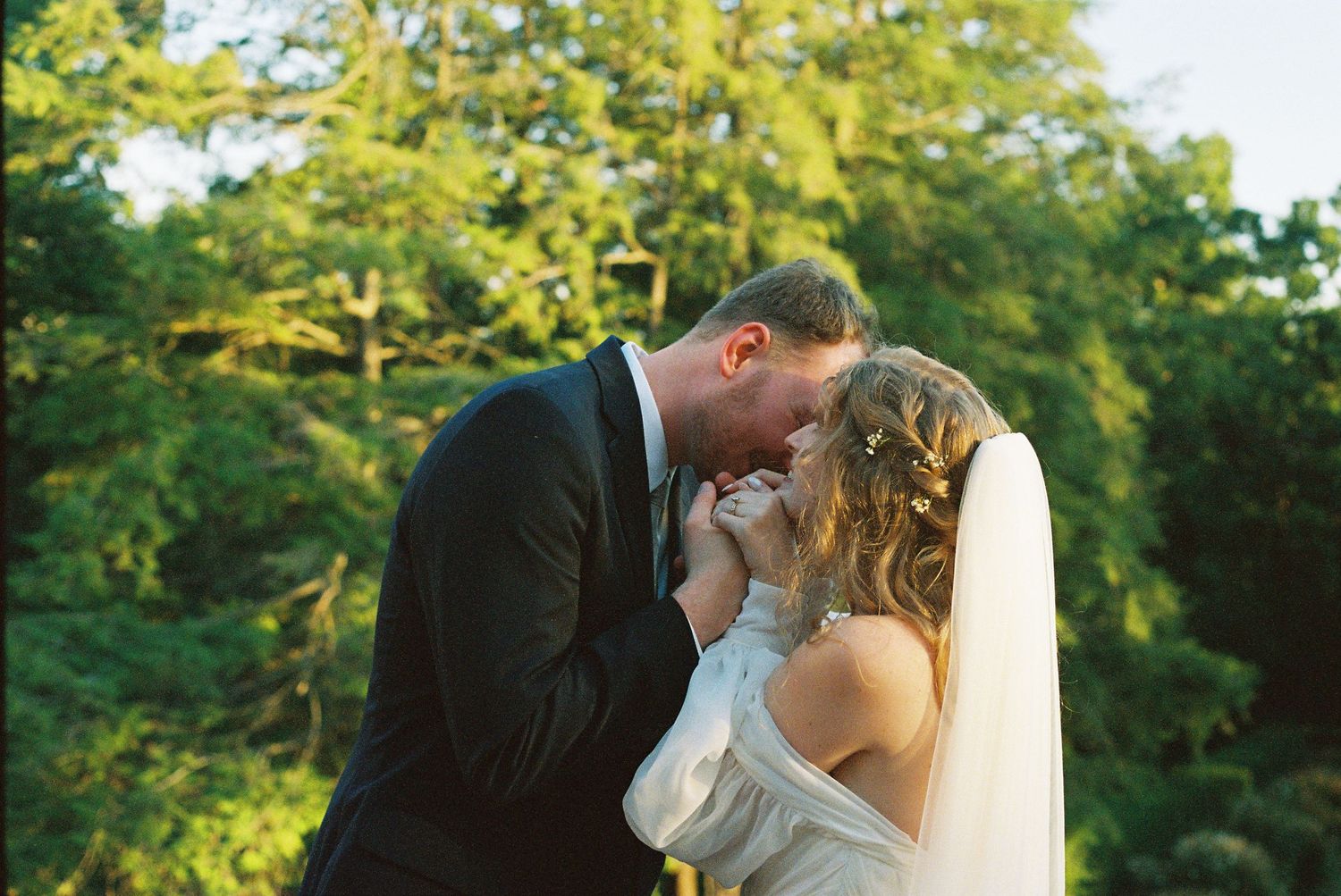 A couple in wedding attire share an intimate moment outdoors with lush green trees in the background.