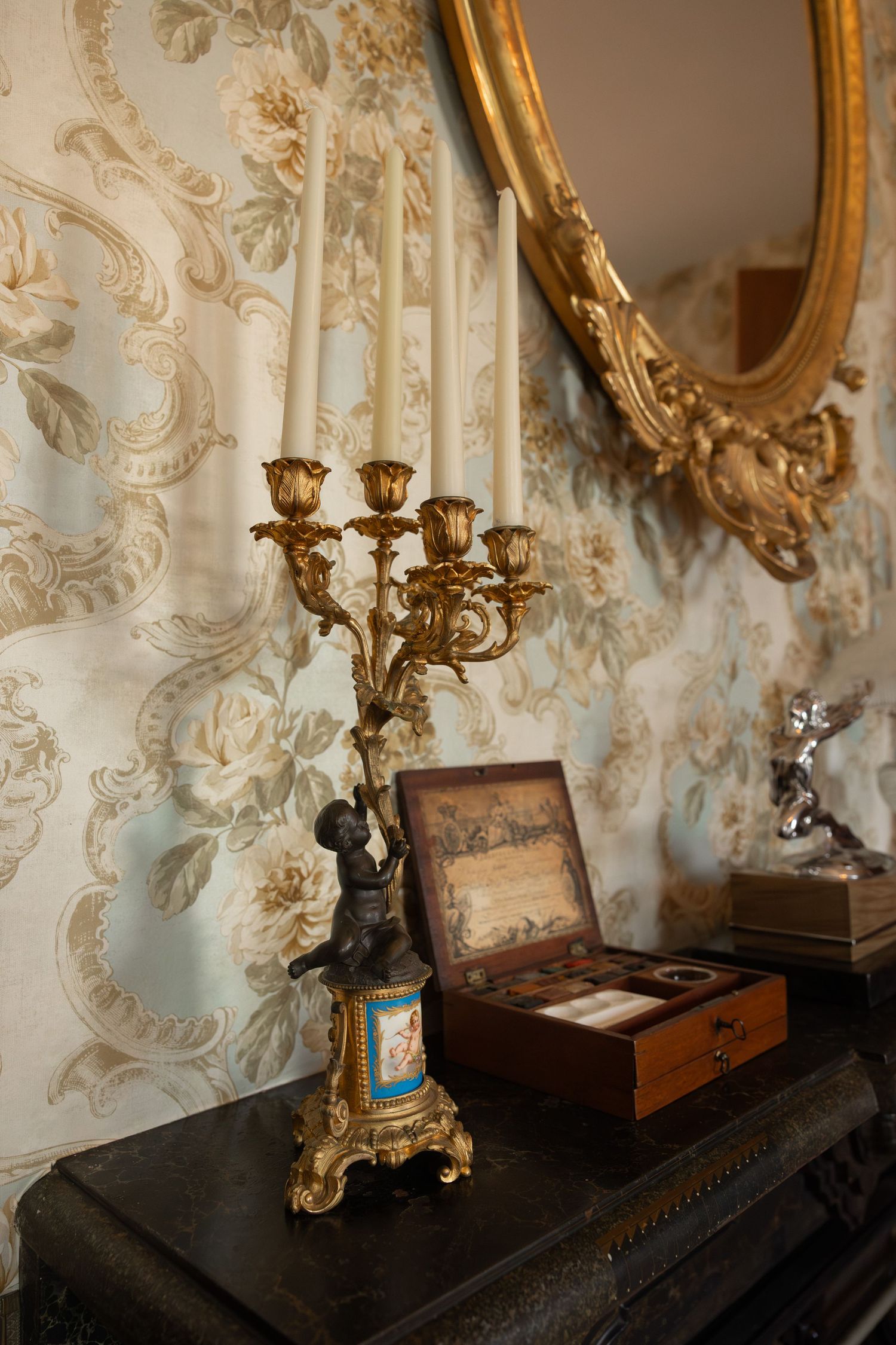 Ornate baroque interior with gold candelabra and decorative mirrors on a wall with intricate damask wallpaper pattern.