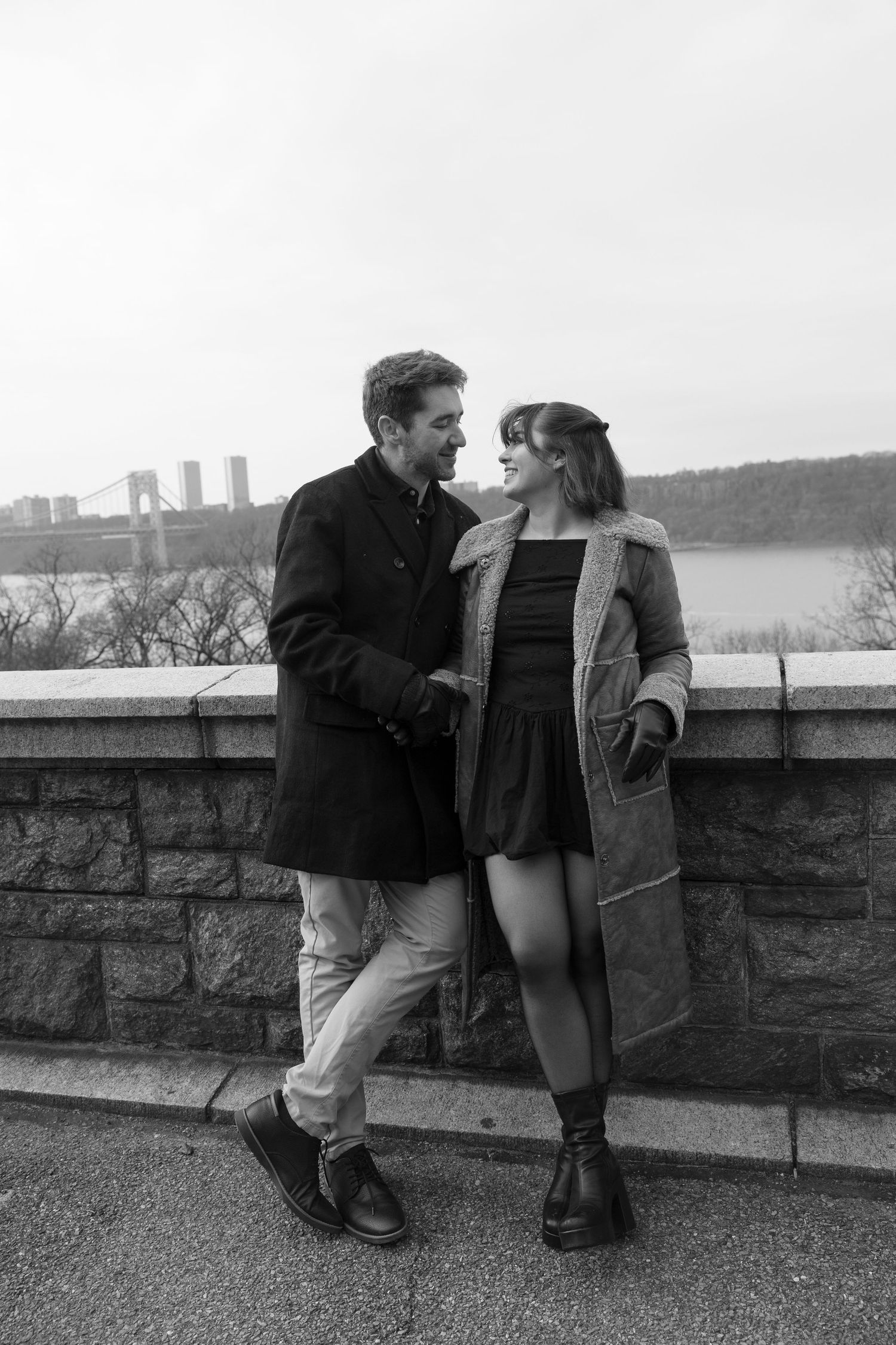 Couple in winter coats sharing an intimate moment along a stone wall overlooking a waterfront.