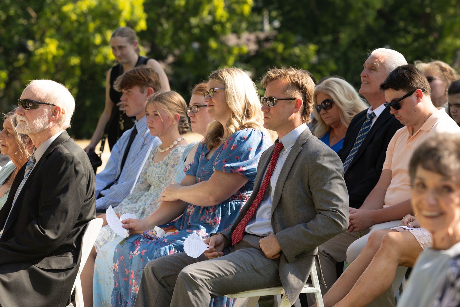 Wedding guests seated outdoors in rows of white chairs during a sunny garden ceremony.