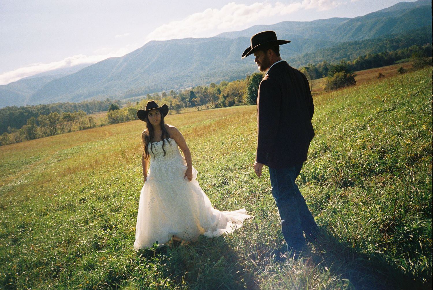 A couple walks through a misty mountain meadow at sunset while holding hands and wearing wedding attire.