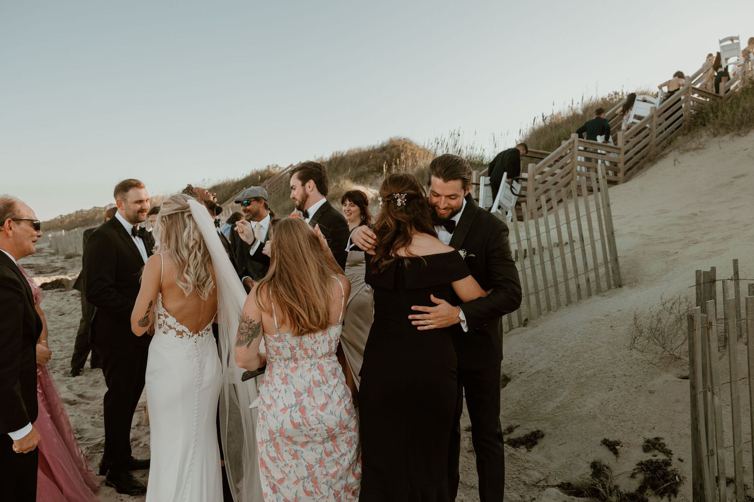 Wedding party members embrace and celebrate on a sandy beach near coastal dunes.