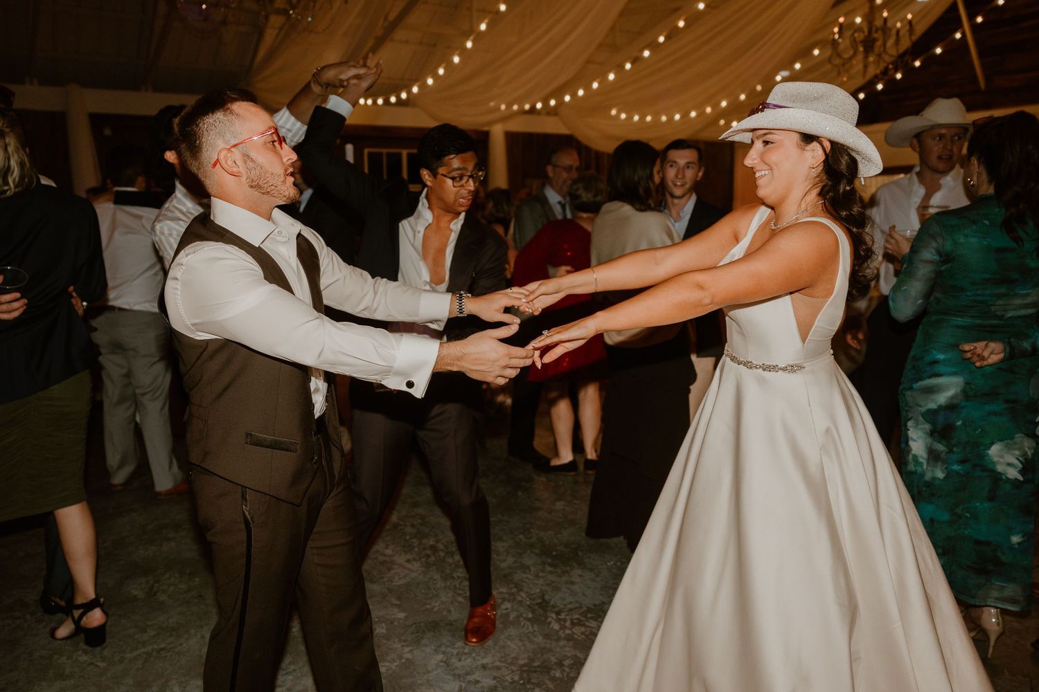 Wedding celebration with guests dancing under string lights in a rustic venue.