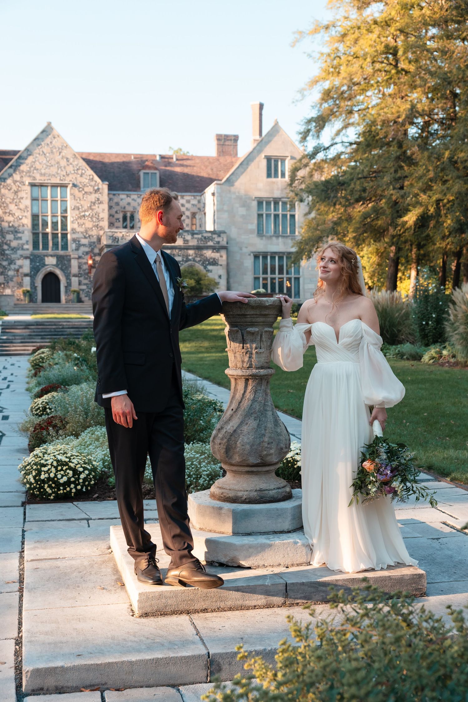 Wedding couple poses for photos outside stone building with garden scenery.